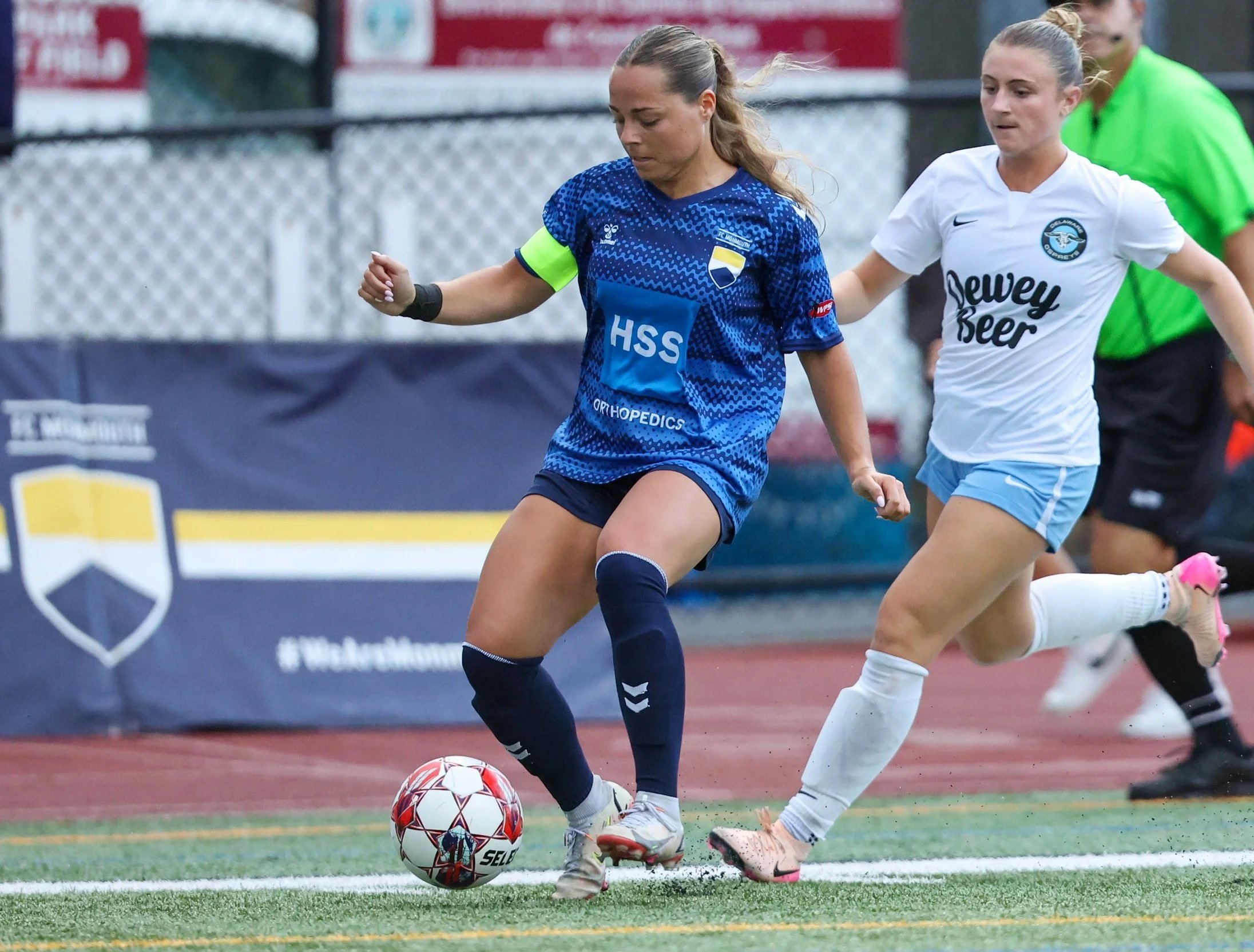 Two female soccer players compete for ball during a match, one in blue and the other in white, with a referee in green in the background.