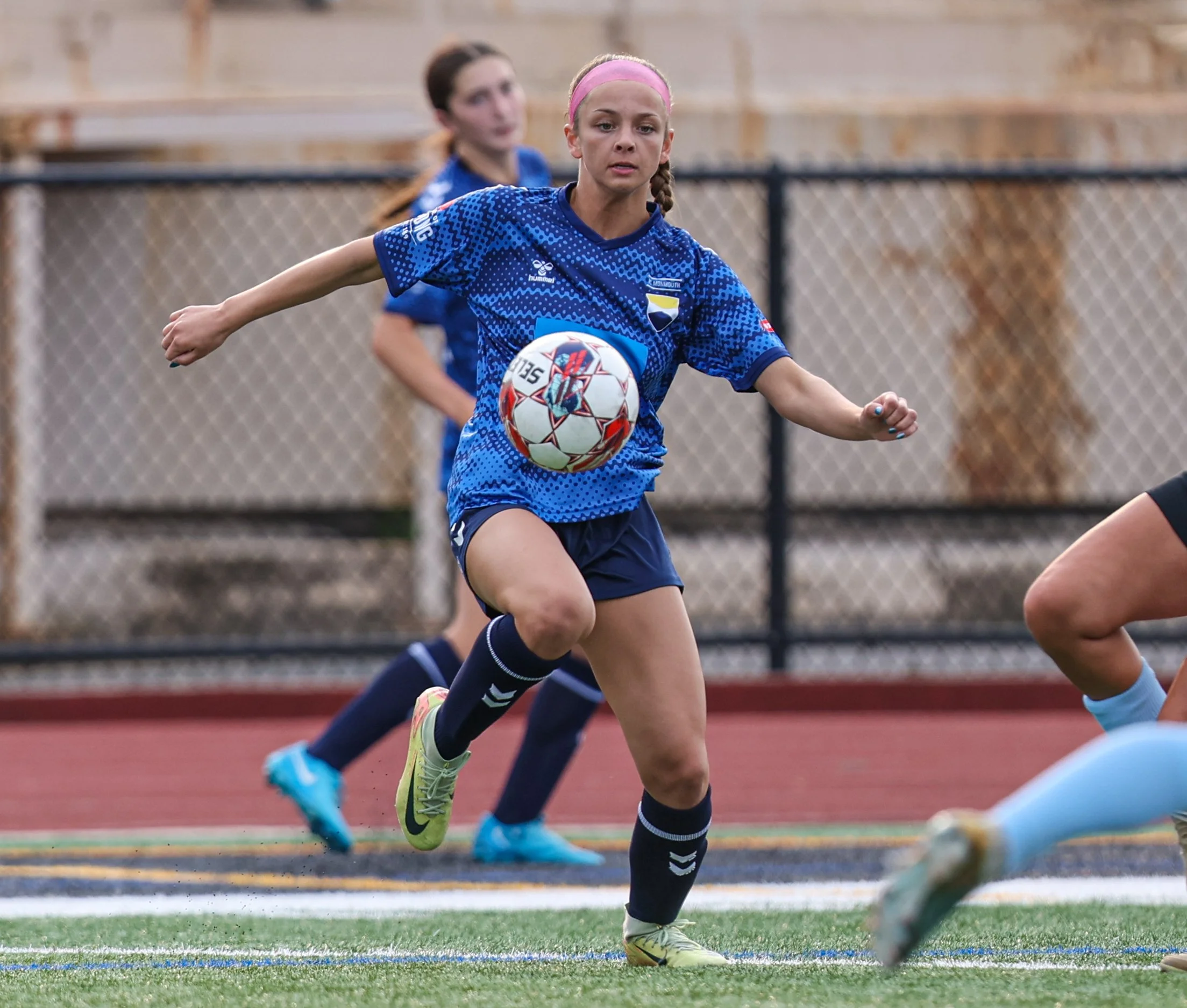 A young girl playing soccer, running with a ball on a field during a game. She is wearing a blue jersey, navy shorts, and neon green cleats, with a pink headband.