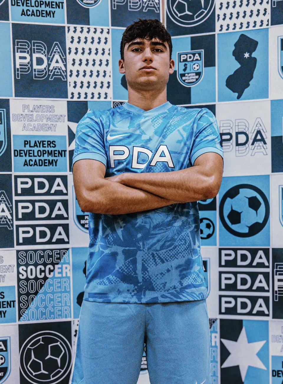 A young man with brown hair running on a soccer field with a soccer ball in front of him. He is wearing a blue jersey with a crest and the letters HSS, blue shorts, and gray shoes. There are trees in the background and another player in a red and white jersey.