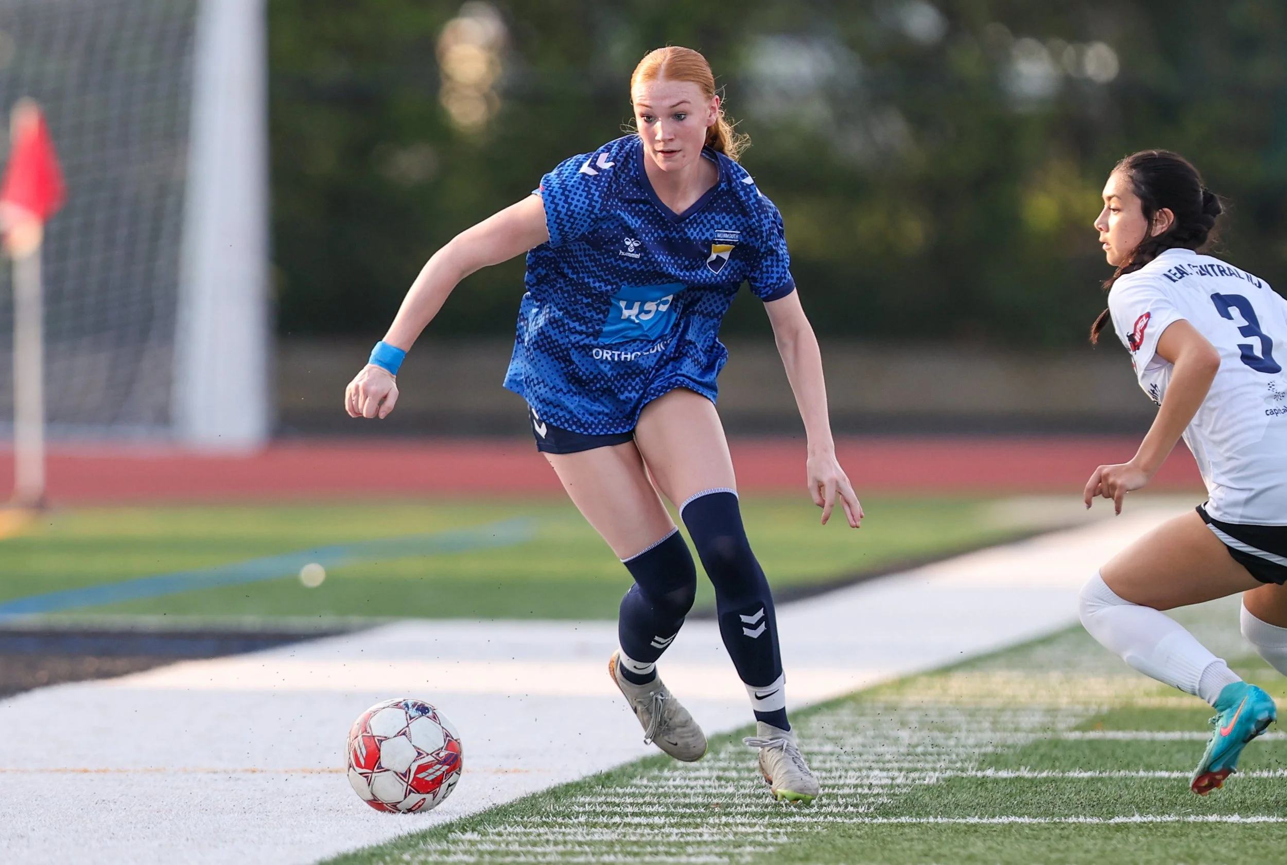 Female soccer players competing for ball on field, one in blue jersey and one in white jersey.