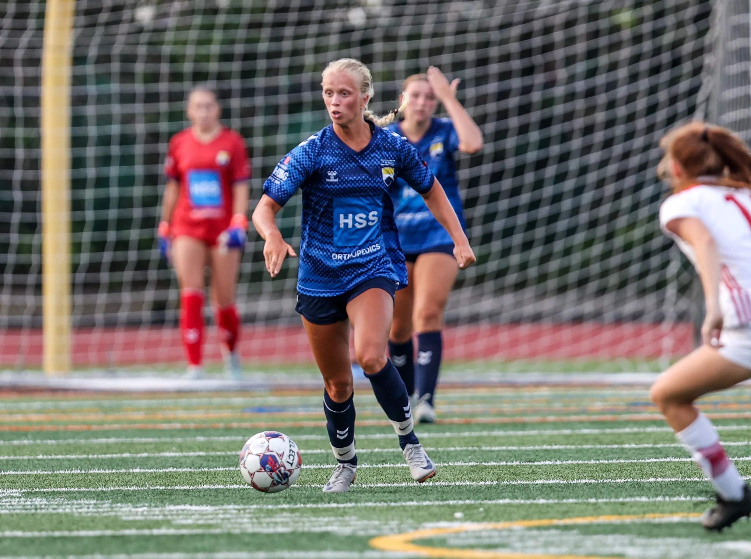 Female soccer player in blue jersey running with the ball towards goal, with defenders and goalkeeper visible in the background on a soccer field.