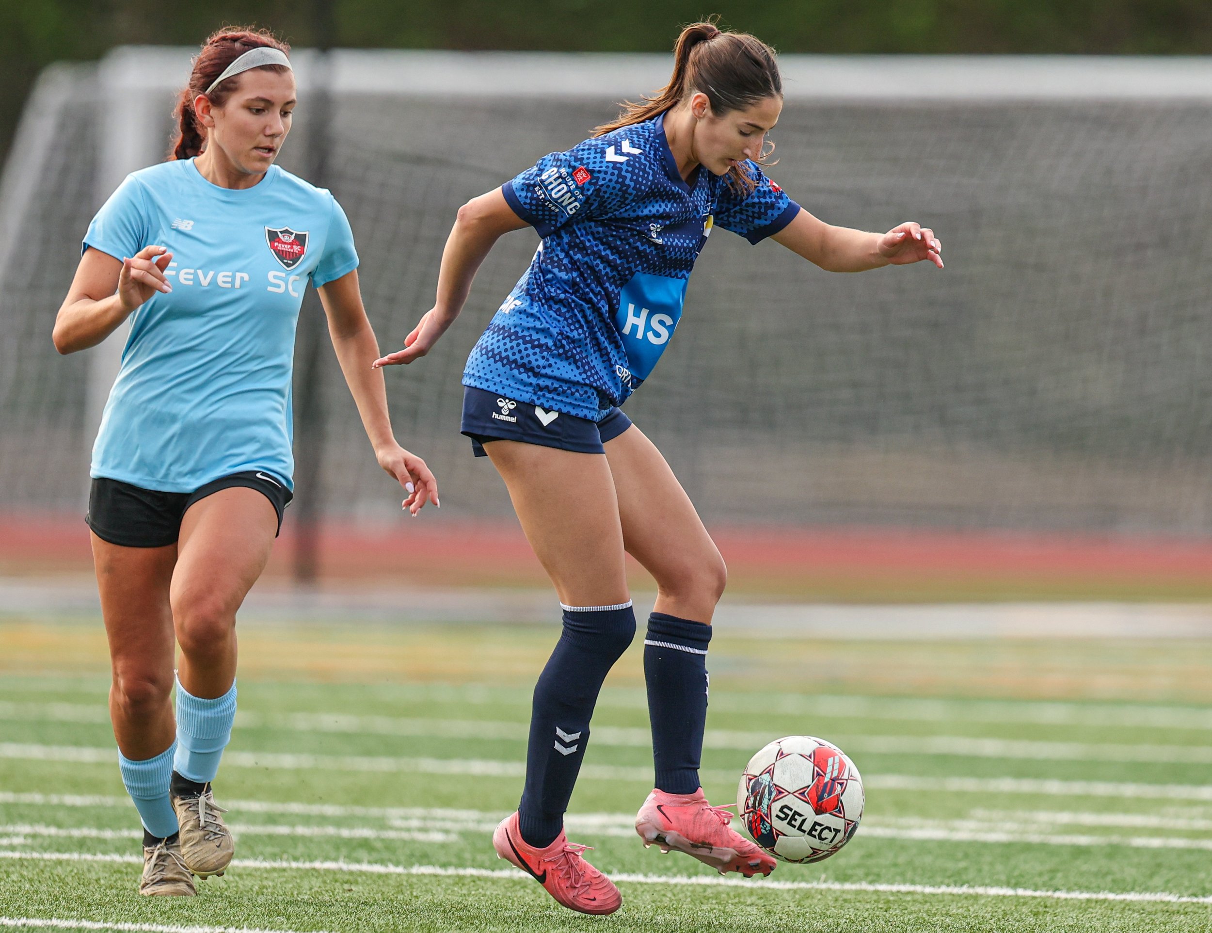 Two women playing soccer on a grass field, one in a blue jersey and the other in a light blue jersey, chasing the ball.