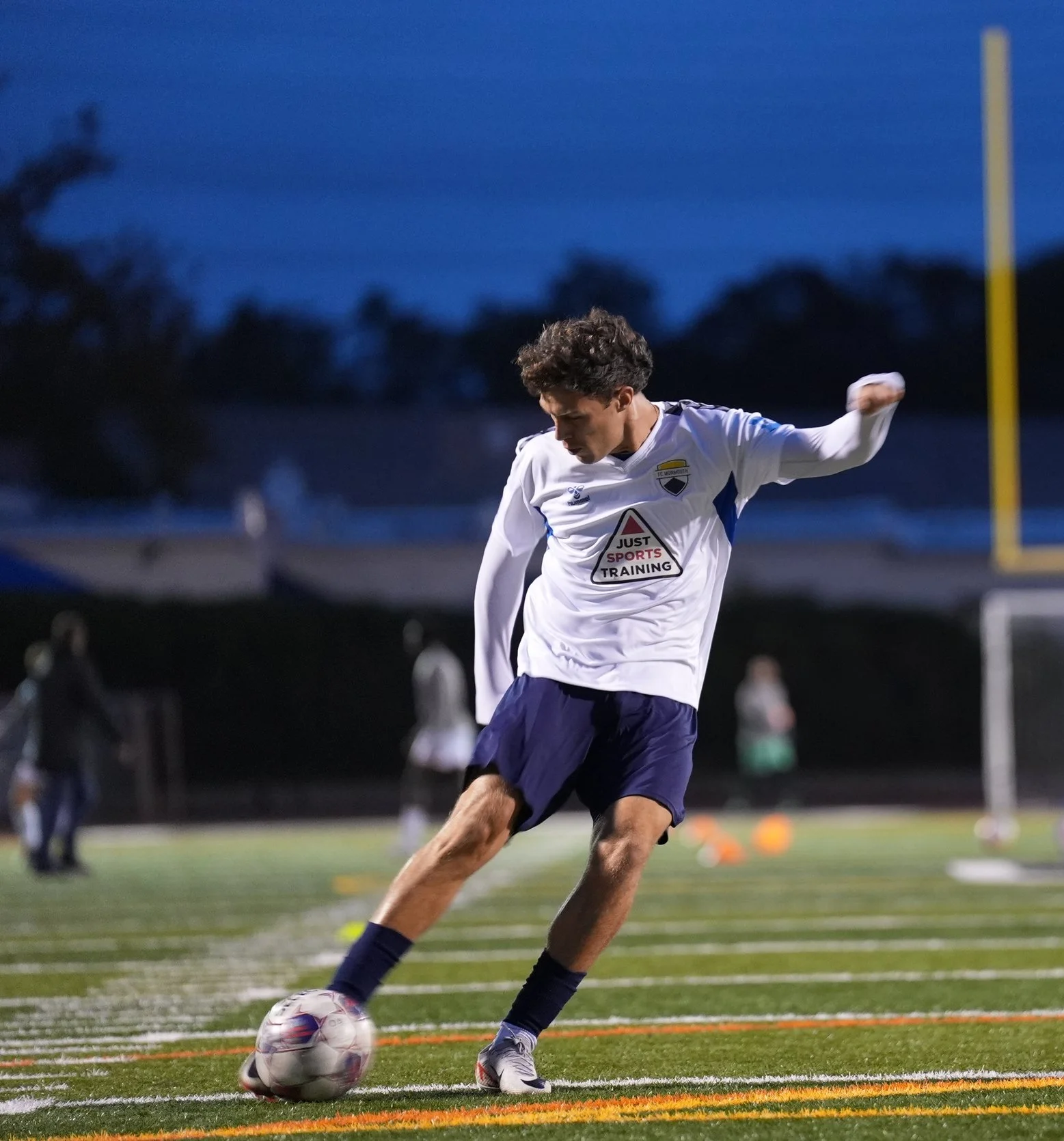 A young man playing soccer on a field during evening, kicking a soccer ball with his right foot, wearing a white jersey and navy shorts.