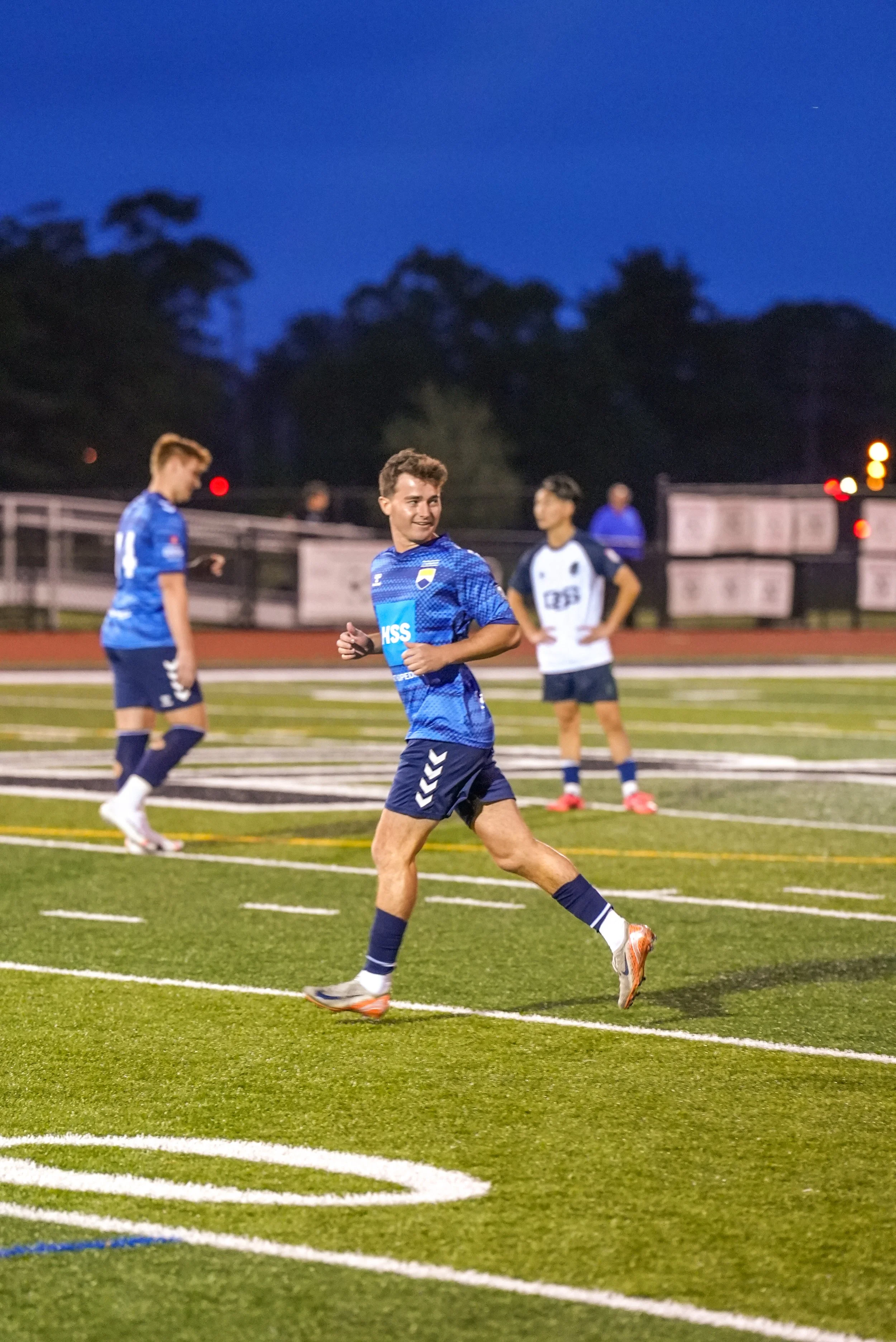 A group of young male soccer players on a field during a game at dusk, with one player in blue jersey running and smiling, and others in the background.