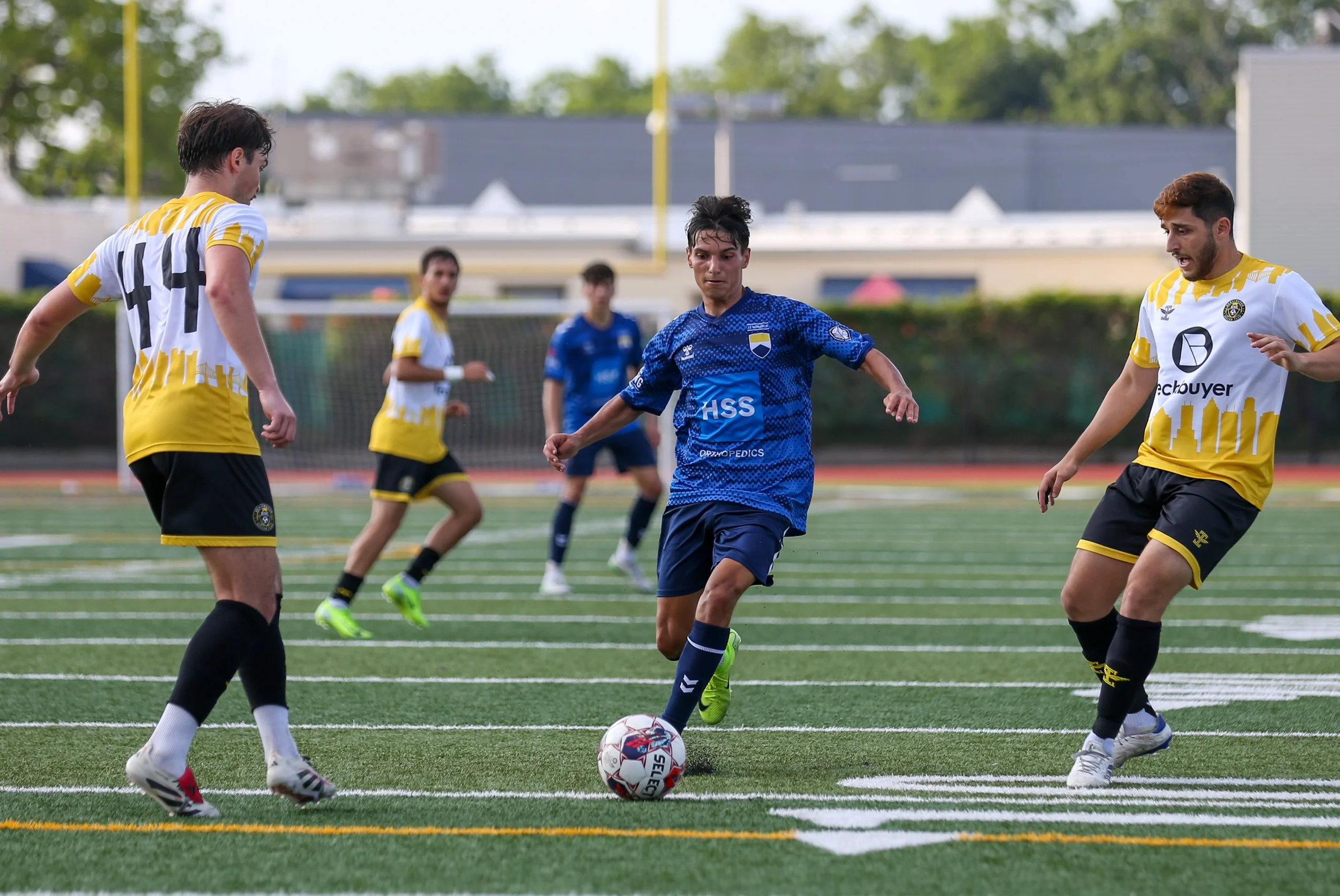 Soccer players in blue and yellow jerseys playing on a field with green turf and white lines during daytime.