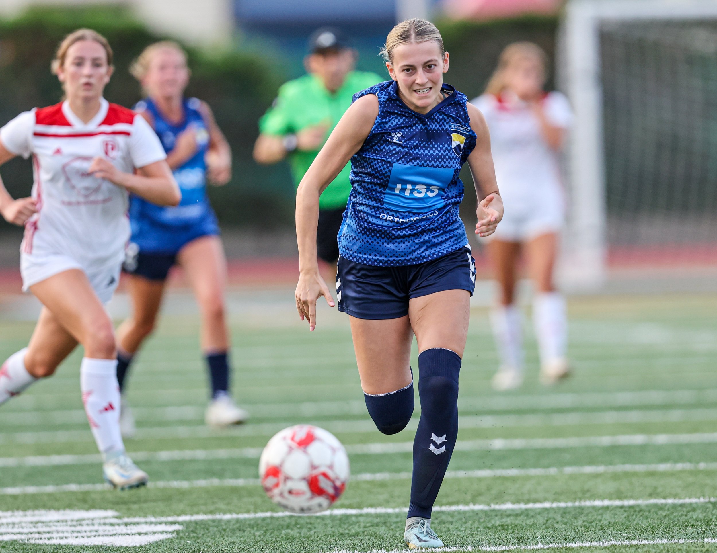 A young woman in a blue sports jersey and shorts running after a soccer ball during a match, with other players and a goal in the background on a field.