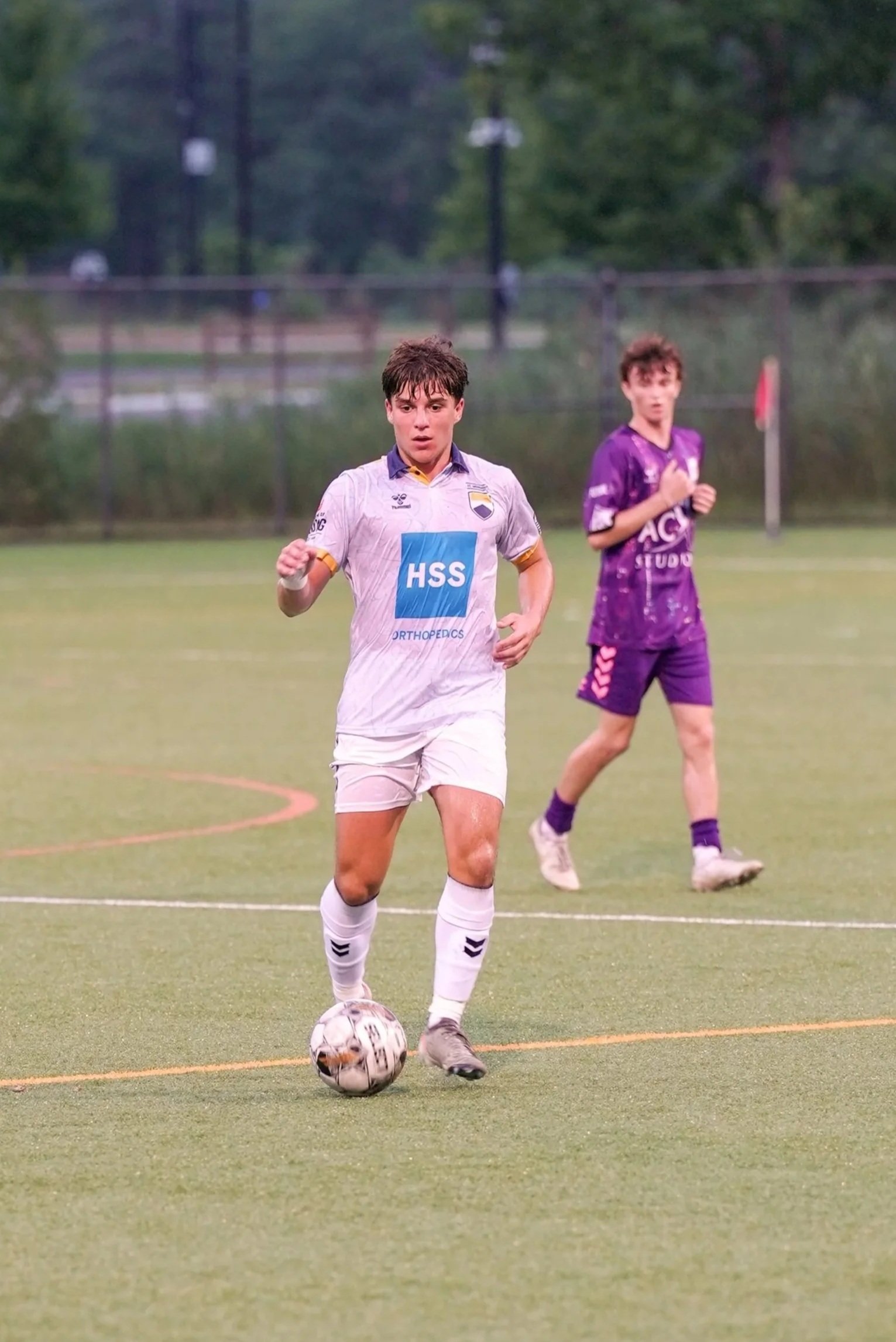 A young male soccer player in a white uniform dribbling a soccer ball on a field, with another player in a purple uniform in the background.