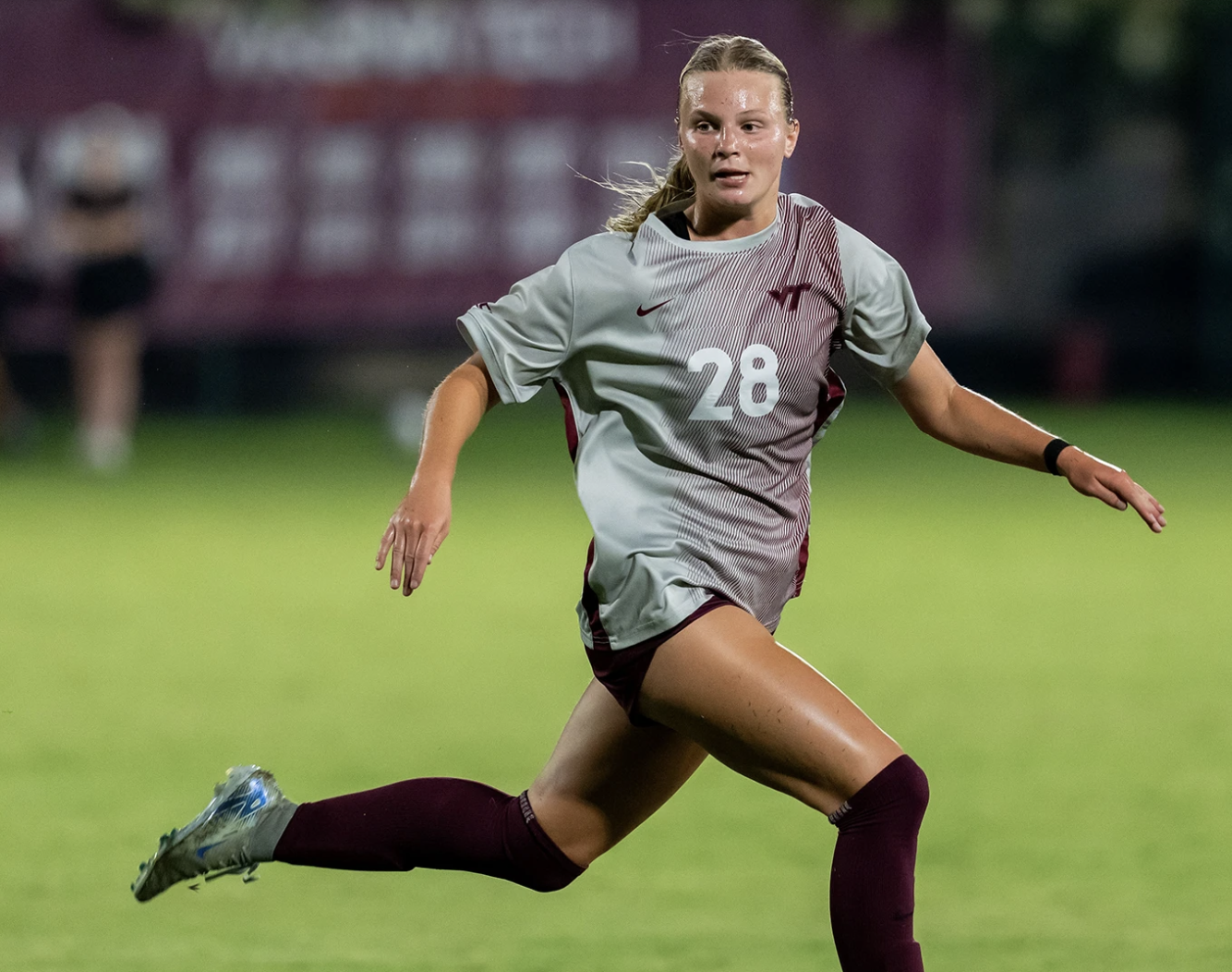 A female soccer player in a white and blue uniform, pink cleats, and long soccer socks standing on a green soccer field during a game.
