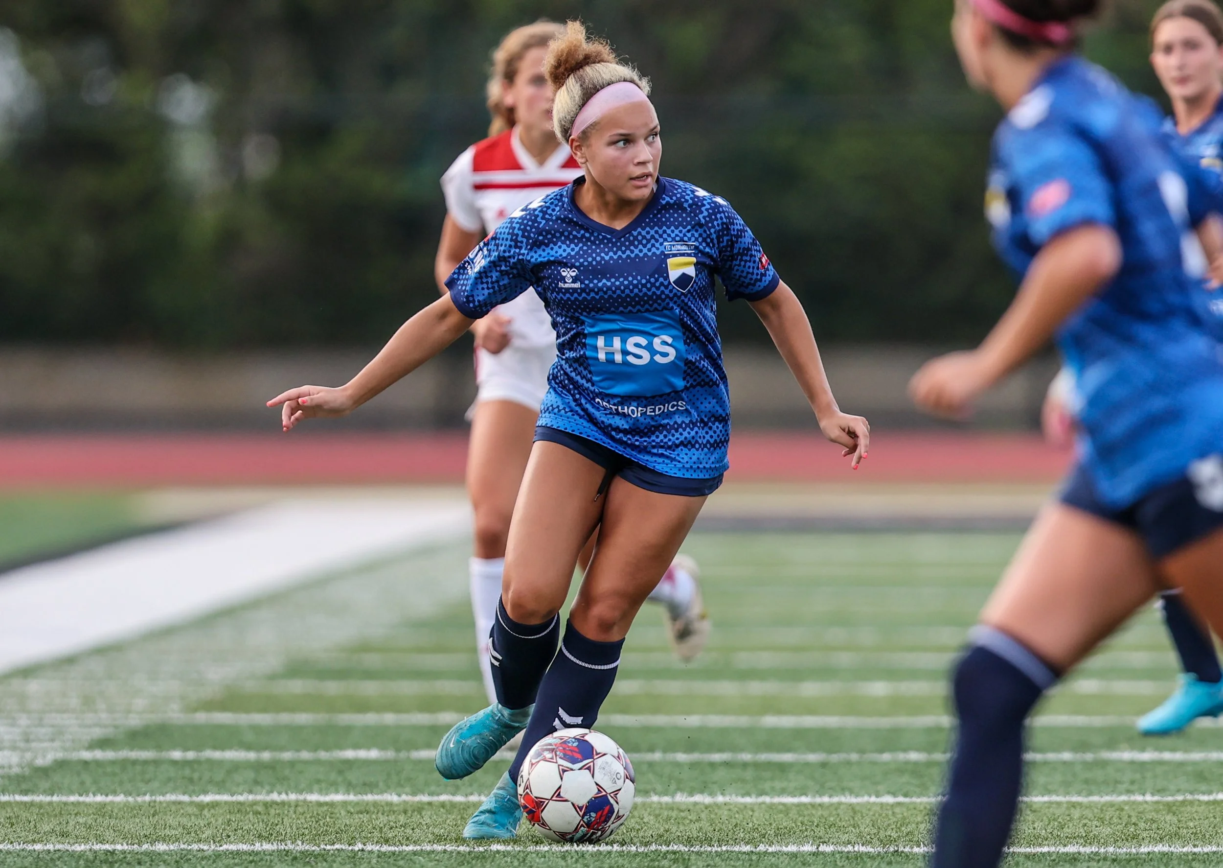 A woman in a blue soccer uniform with a pink headband, holding a soccer ball, on a field with other women in the background during a game.