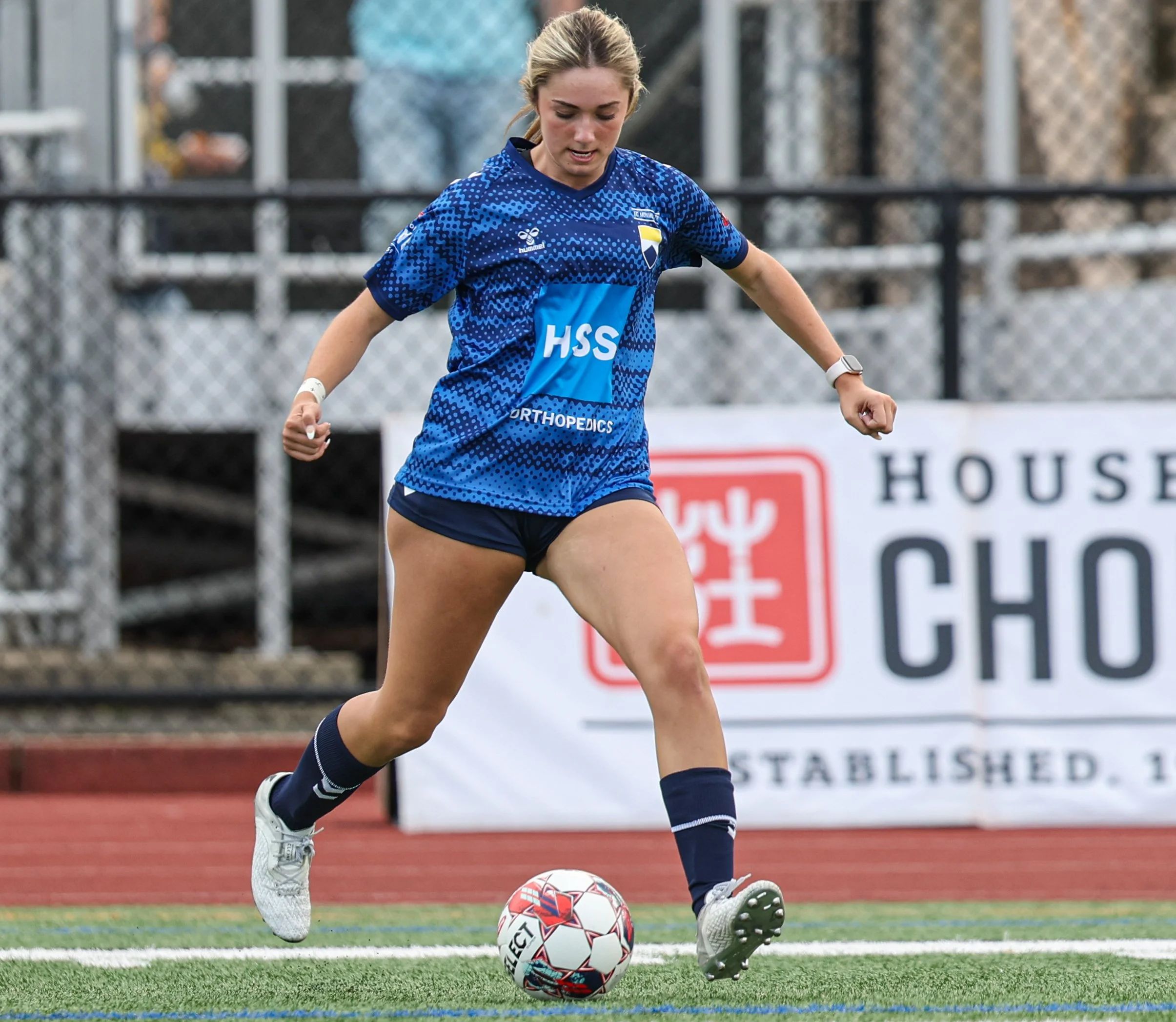 A female soccer player in a blue jersey and shorts kicking a soccer ball on a field during a game.