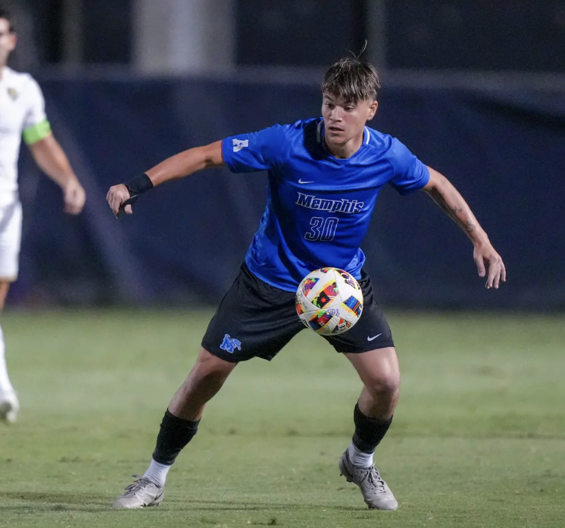 A soccer player in a white and blue uniform on the field at night with other players in the background.