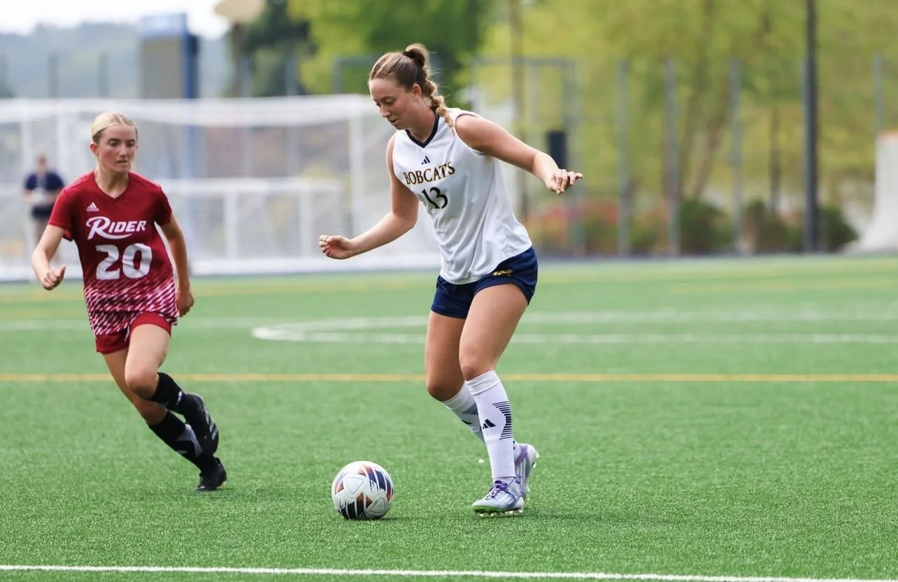 Female soccer player in white uniform with pink cleats chasing a soccer ball on a green field during a game, with a goalkeeper in black in front of the goal in the background.