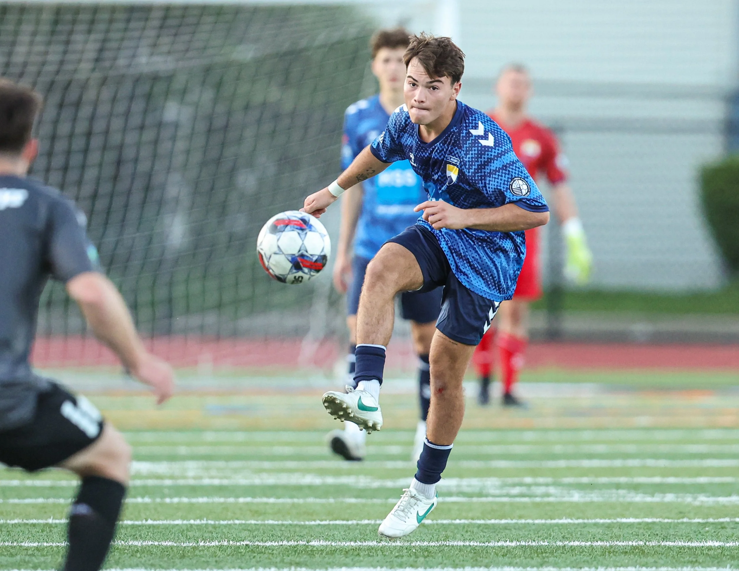 A soccer player in a blue jersey kicks a soccer ball on a field during a match, with other players and a coach in the background.