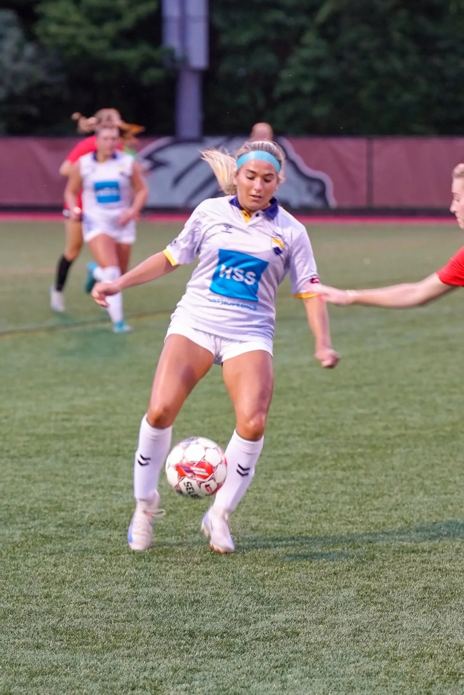 A female soccer player in a white uniform with a blue headband and sponsor logo, kicking a soccer ball on a field, with other players and trees in the background.