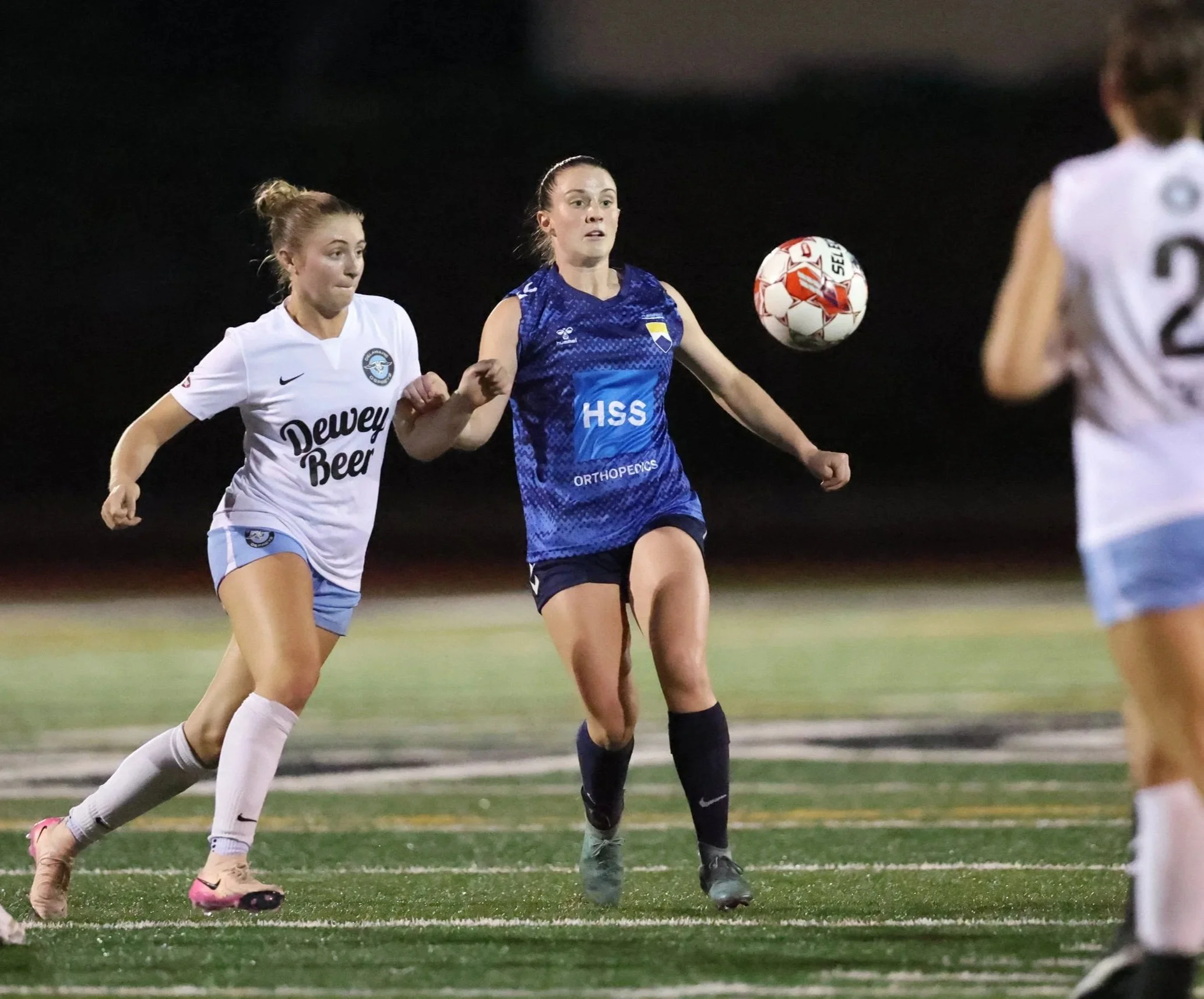 Soccer players, one in a white jersey and one in a blue jersey, compete for the ball on a night game