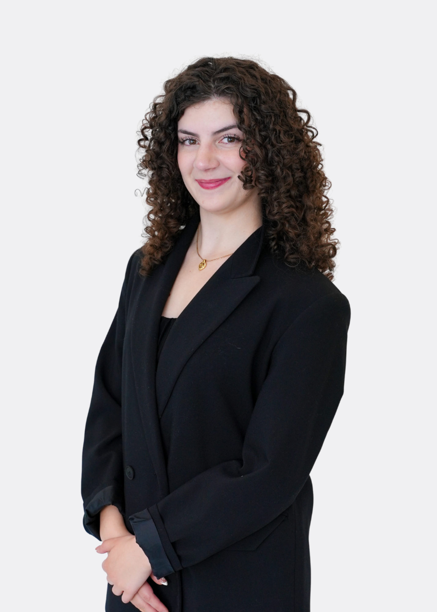 Professional headshot of a woman with straight, shoulder-length red hair wearing a black blazer and black top. She is smiling warmly, accessorized with layered necklaces, rings, and bracelets, standing against a light gray background.