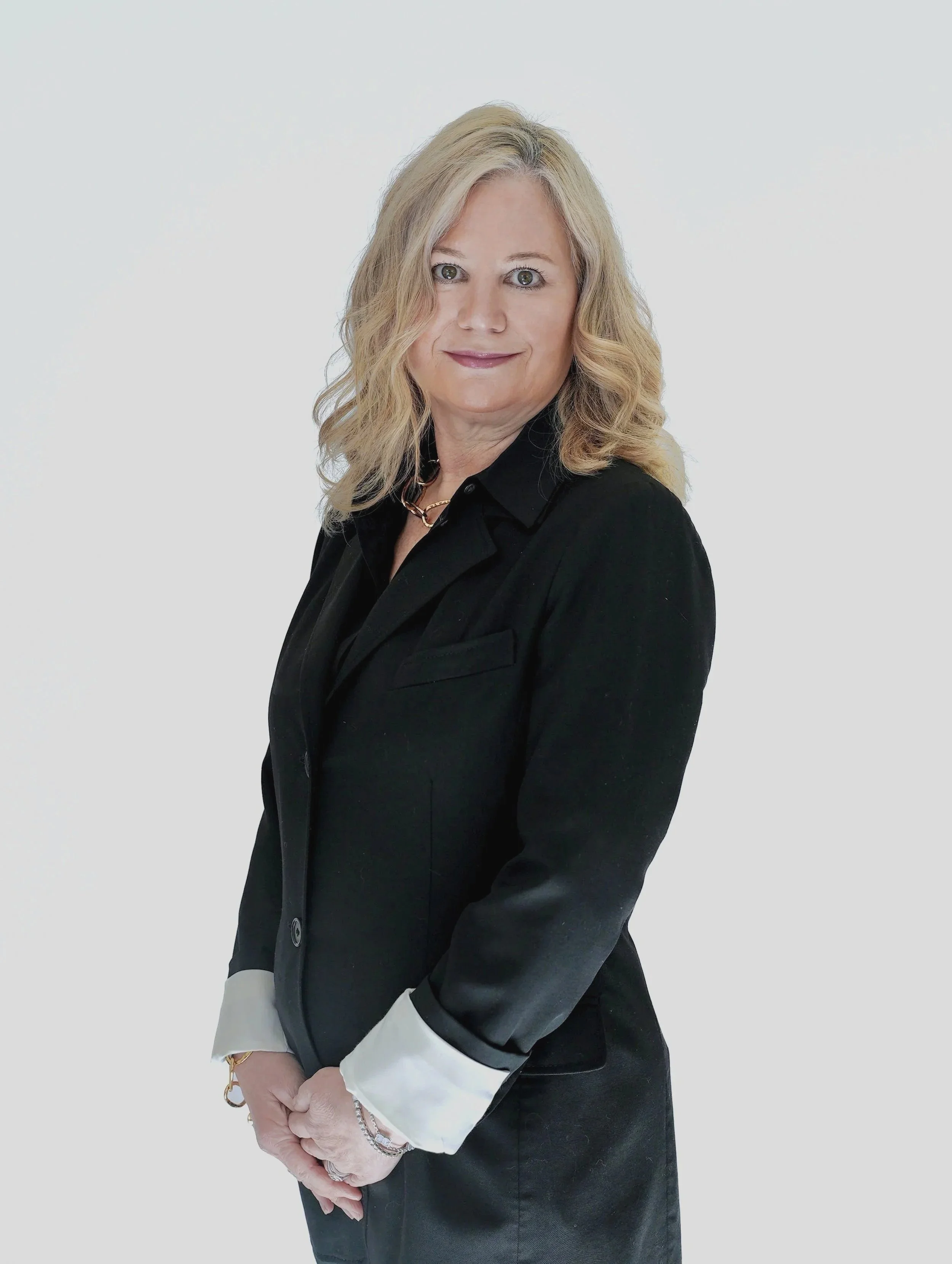 Professional headshot of a person with long, straight dark hair styled half-up with a decorative hairpin. They are wearing a black suit with silver safety pin accents and a black shirt and tie. Standing against a light gray background.
