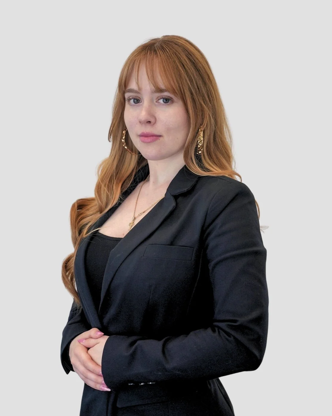 Professional headshot of a woman with blonde, shoulder-length wavy hair, wearing a sleeveless black dress. She is standing against a light gray background with a calm, friendly expression and her hands gently clasped in front of her.