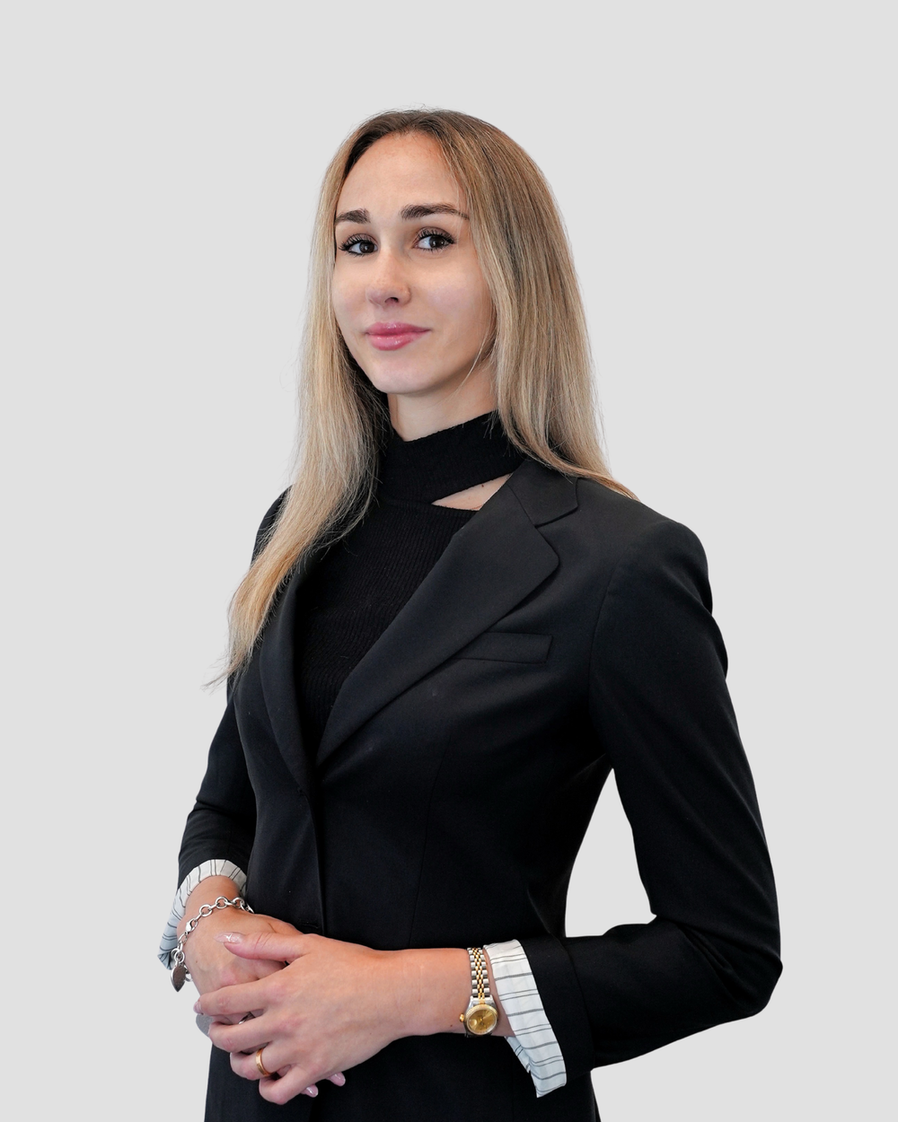 Professional headshot of a woman with straight, light blonde hair wearing a black blazer with white pinstriped cuffs and a black top with a cutout neckline. She is standing against a light gray background with a confident expression.