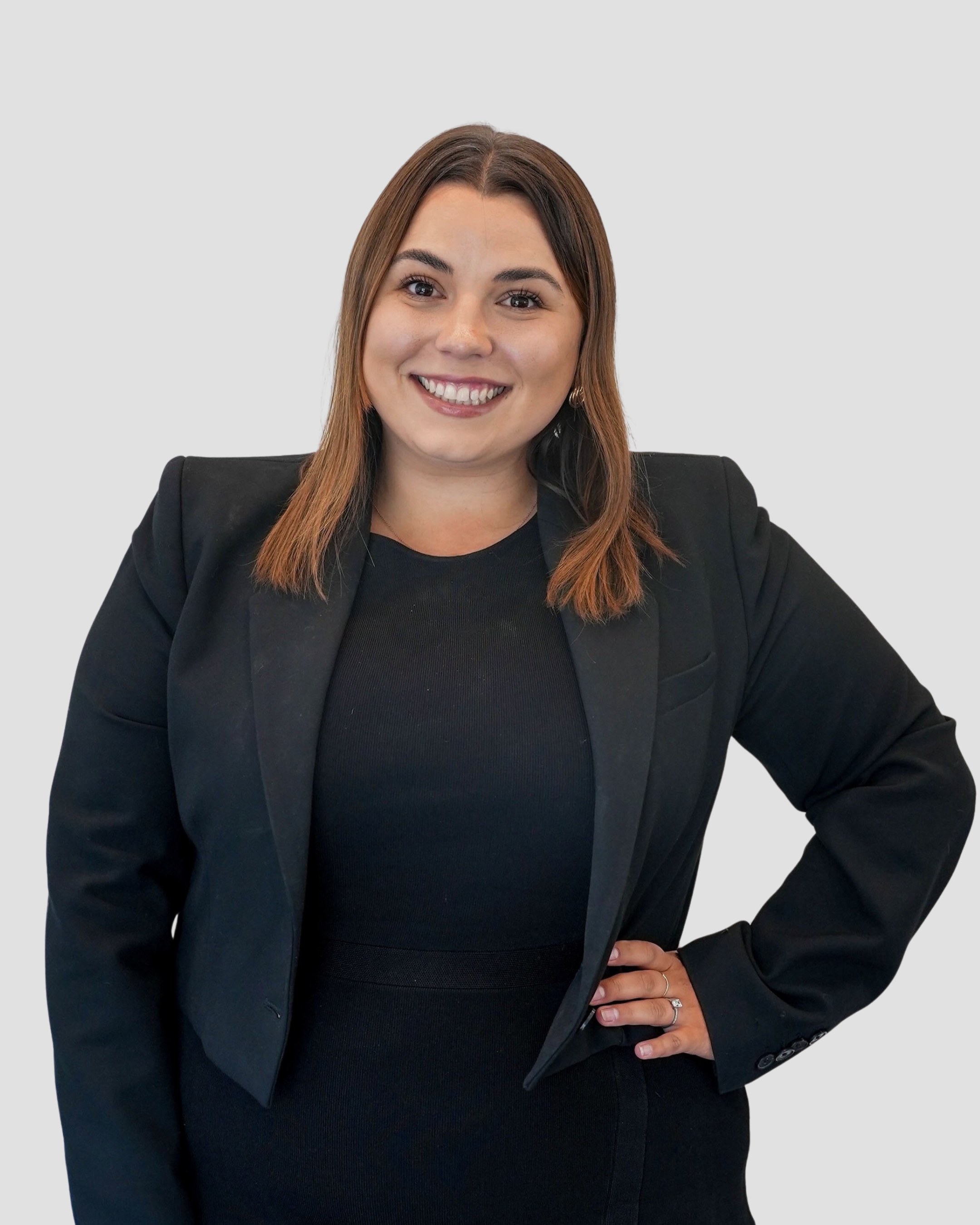 Professional headshot of a woman with straight, shoulder-length brown hair, wearing a black blazer over a black top. She is smiling warmly, standing against a plain light gray background with one hand on her hip in a confident and approachable pose.