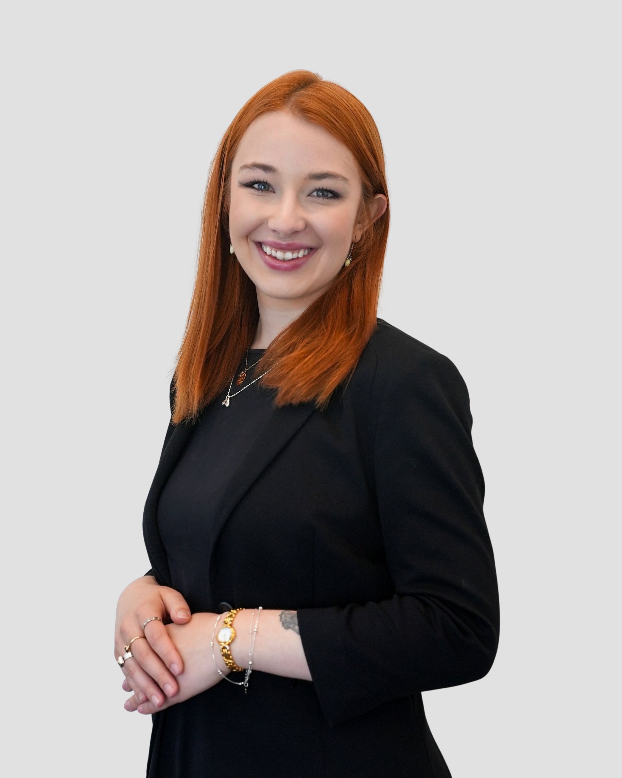 Professional headshot of a woman with straight, shoulder-length brown hair, wearing a black blazer over a black top. She is smiling warmly, standing against a plain light gray background with one hand on her hip in a confident and approachable pose.