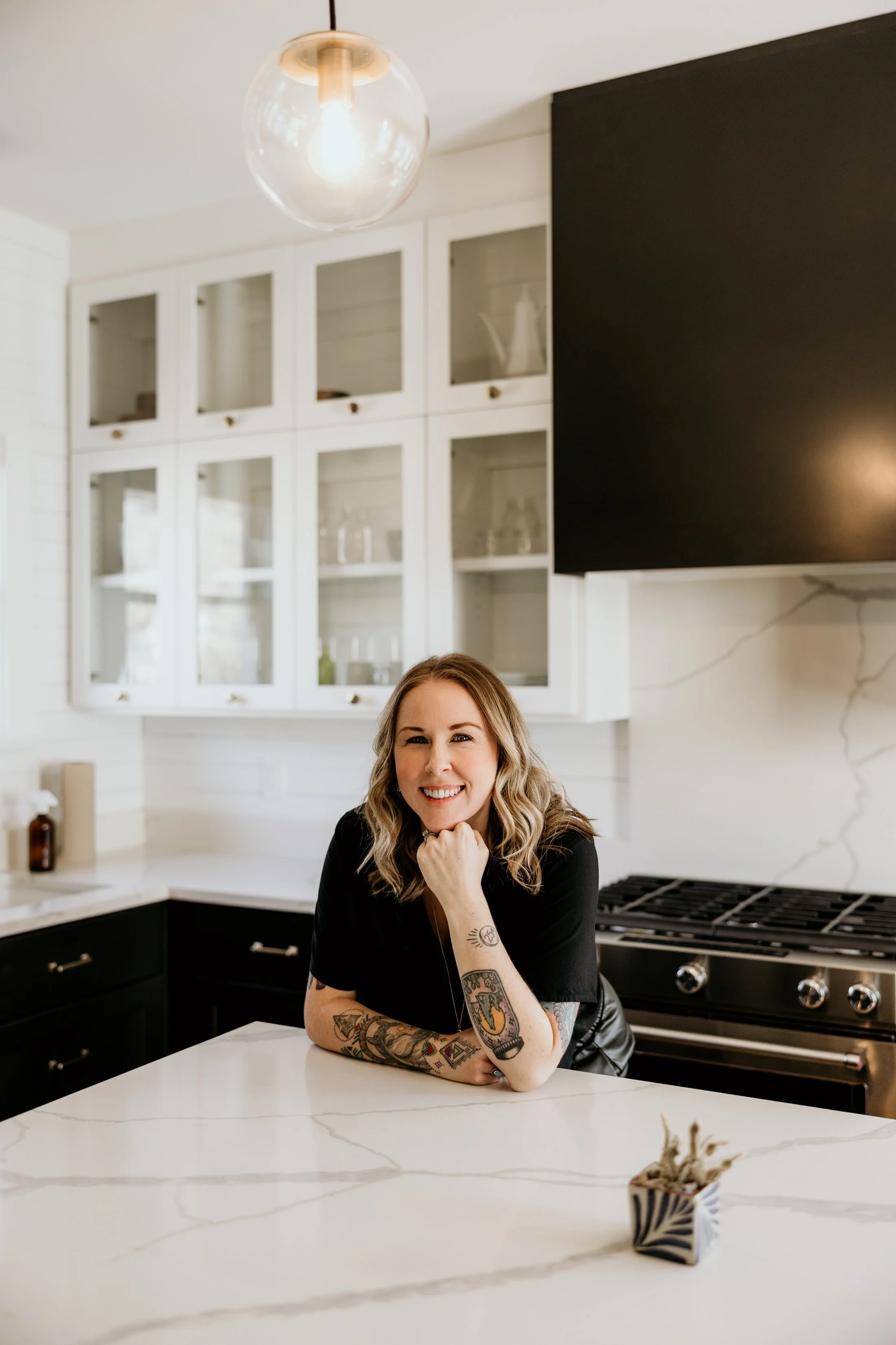 A woman with tattoos and blonde hair smiling at a kitchen counter with white marble countertop, black cabinets, and a white decorative box.