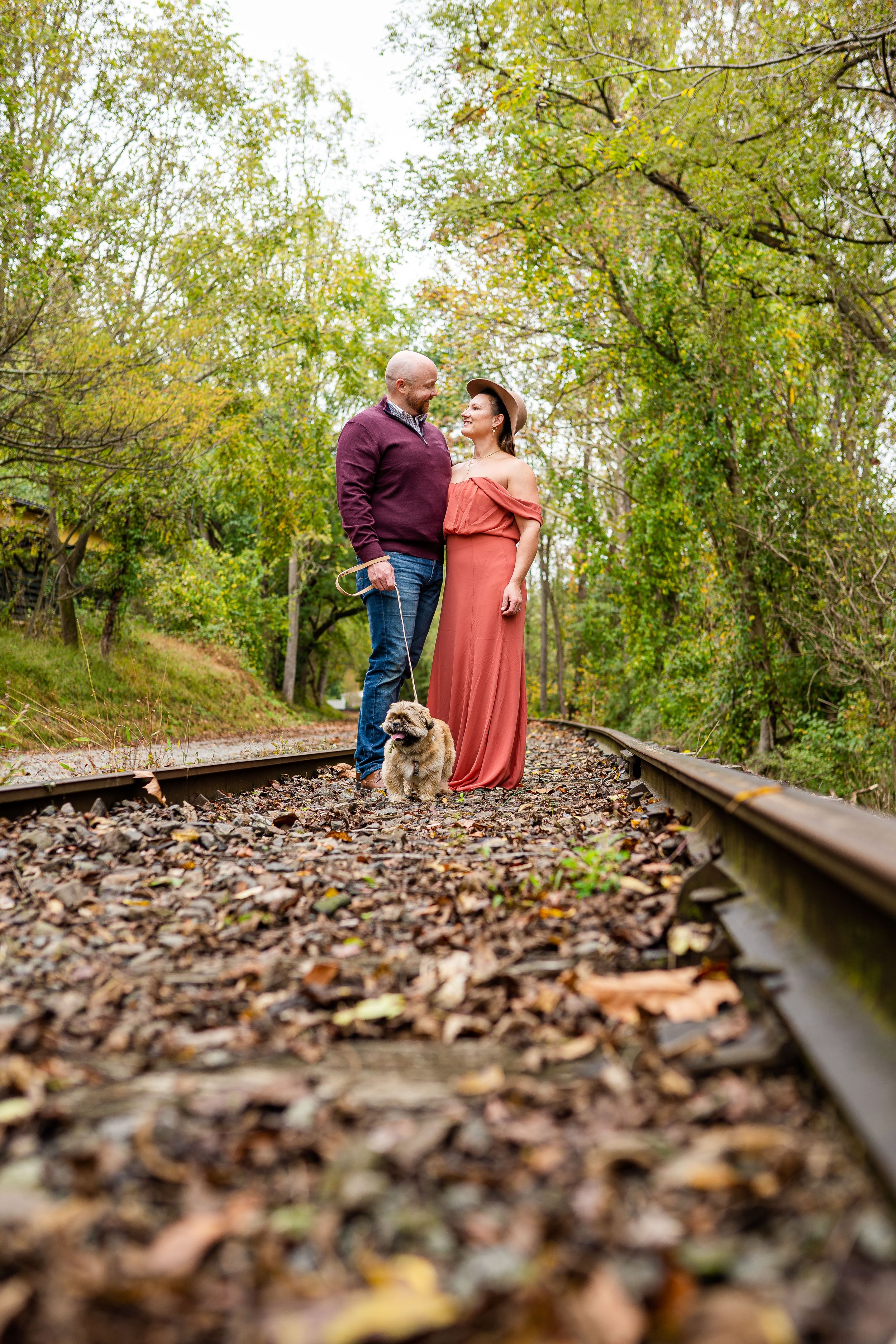 An engaged couple and they small dog smile at each other along a rail trail in Dallastown PA