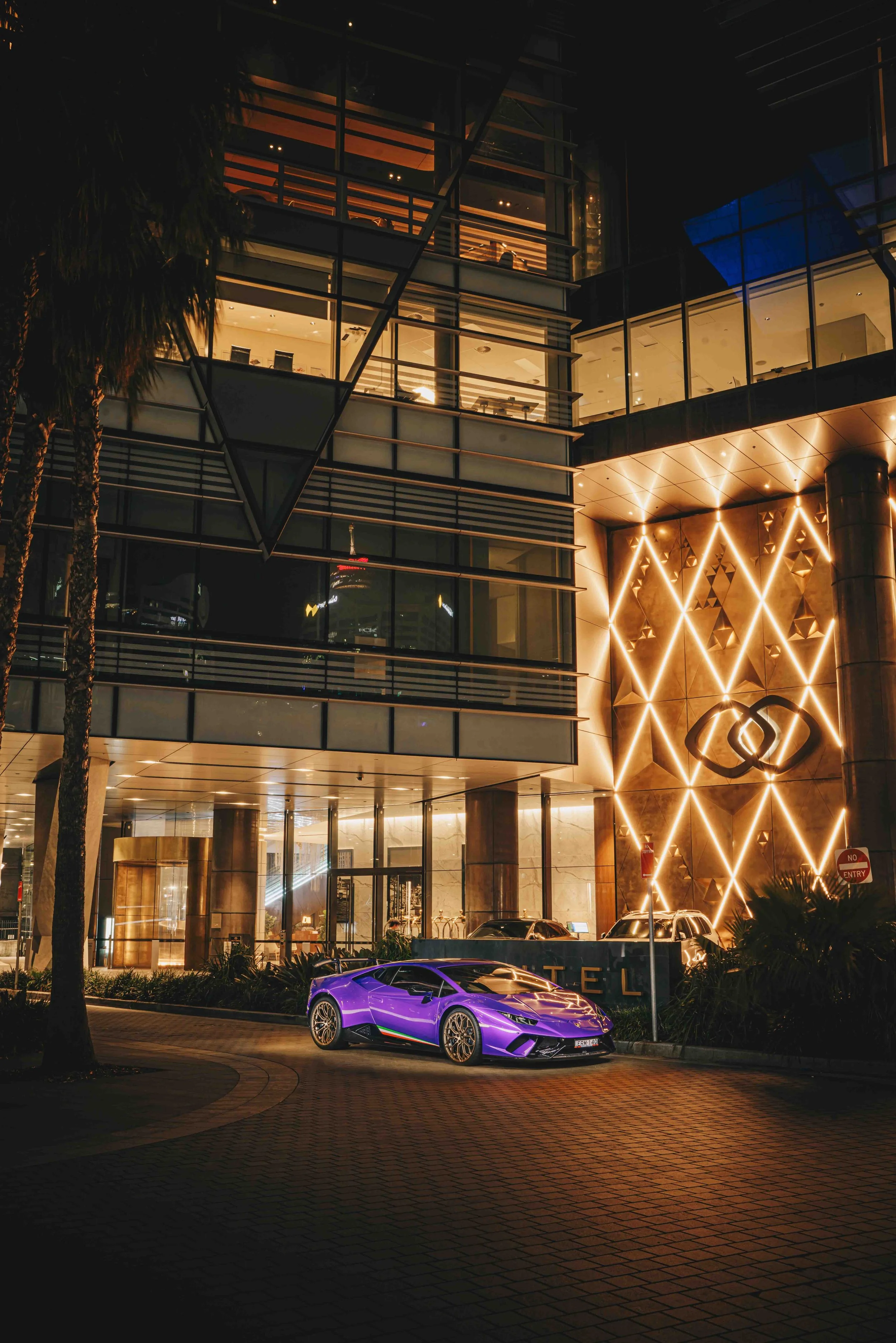 Night view of a modern building with illuminated geometric decoration, tall glass windows, and a purple Lamborghini sports car parked in front.