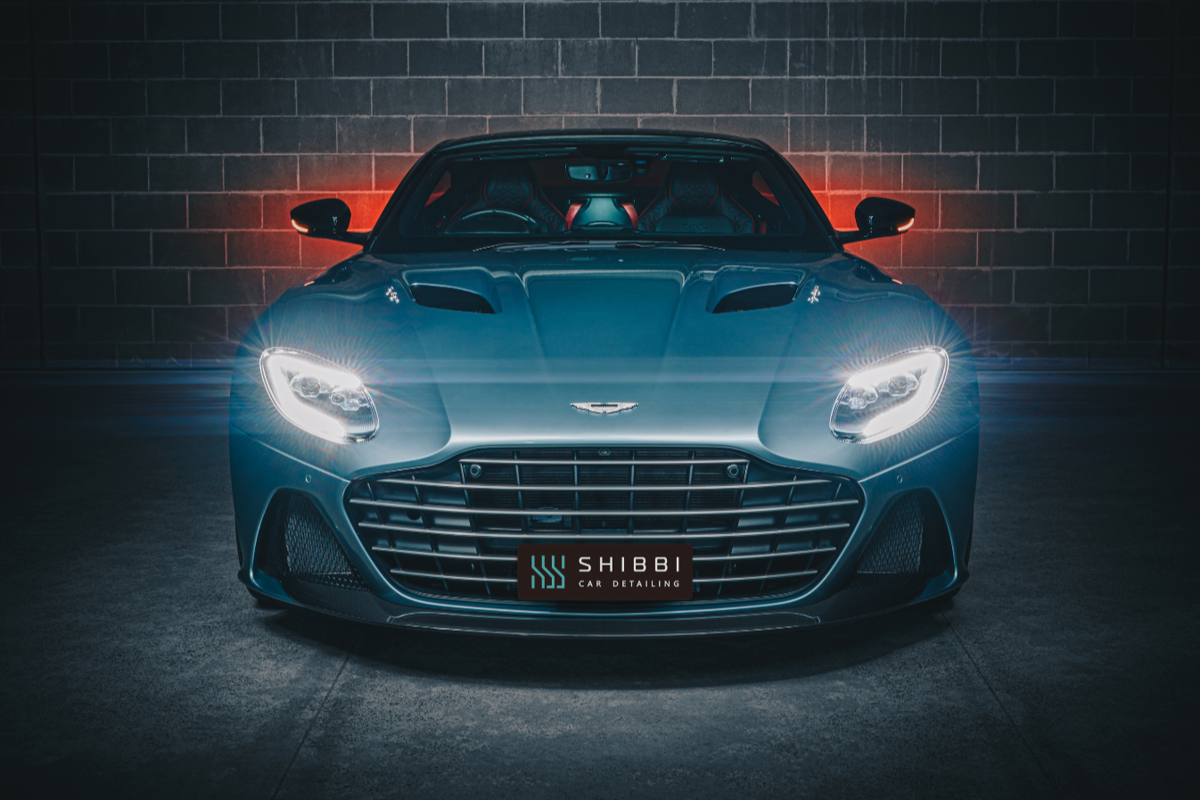 A sleek blue Aston Martin sports car with illuminated headlights parked in front of a brick wall, featuring a Shibbi Car Detailing license plate.