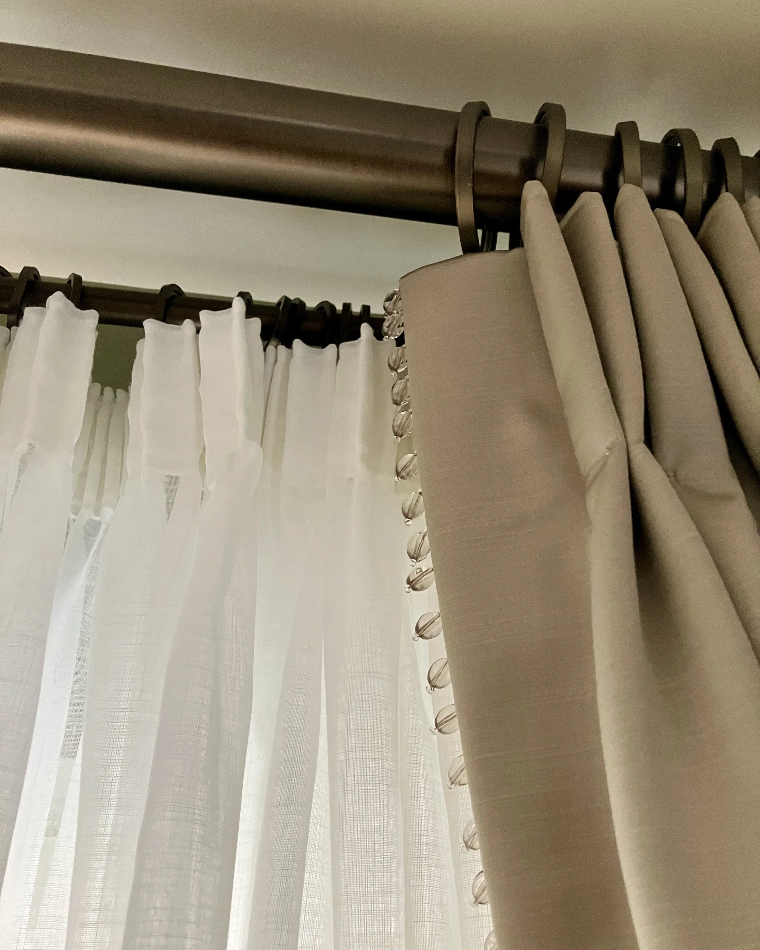 Close-up of a window with sheer white curtains and beige drapes, hung on dark curtain rods with decorative finials.