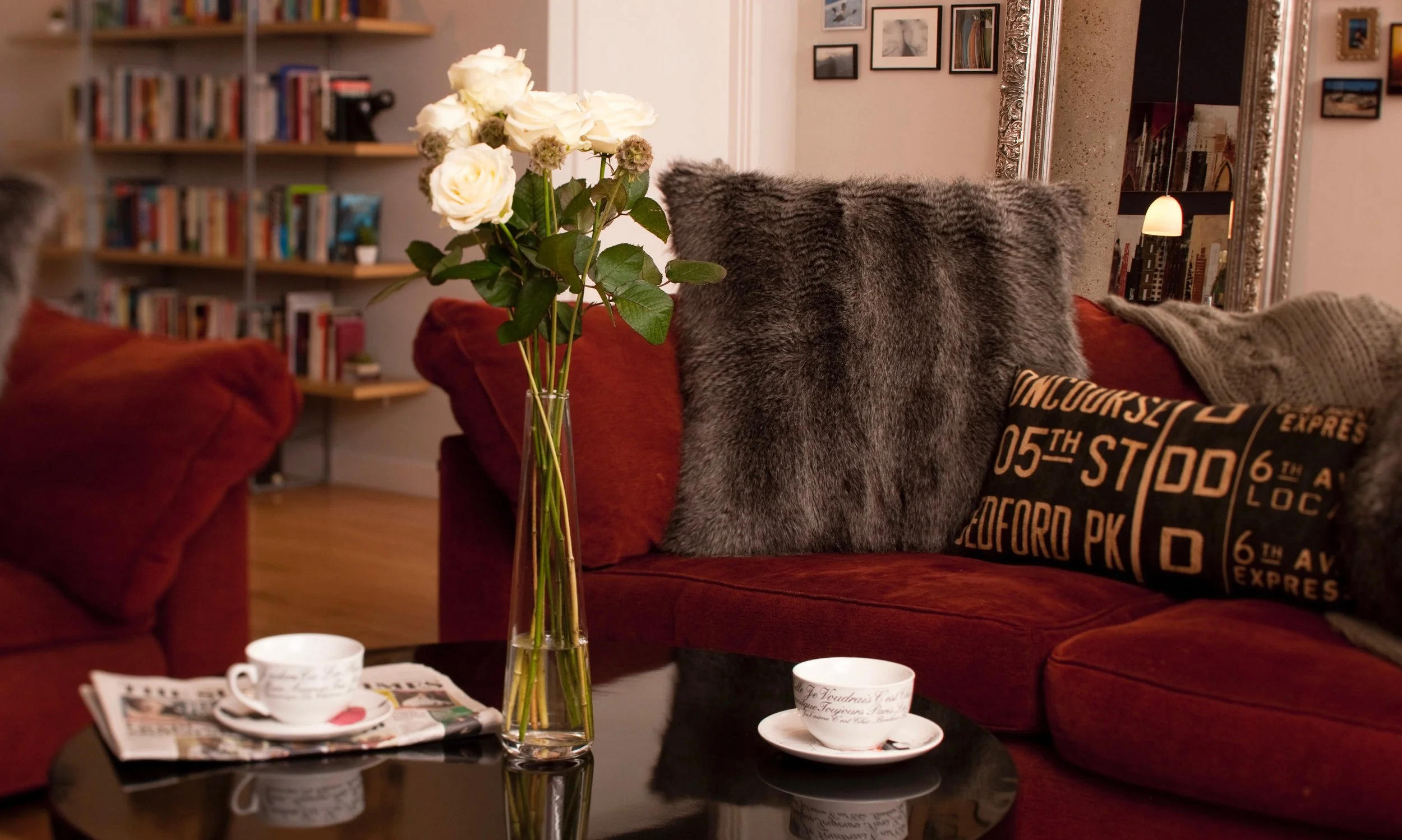 A cozy living room with a terracotta damask sofa, a vase of white roses on a black coffee table, and photos on the wall. A fuzzy grey blanket and a pillow with city names are on the sofa.