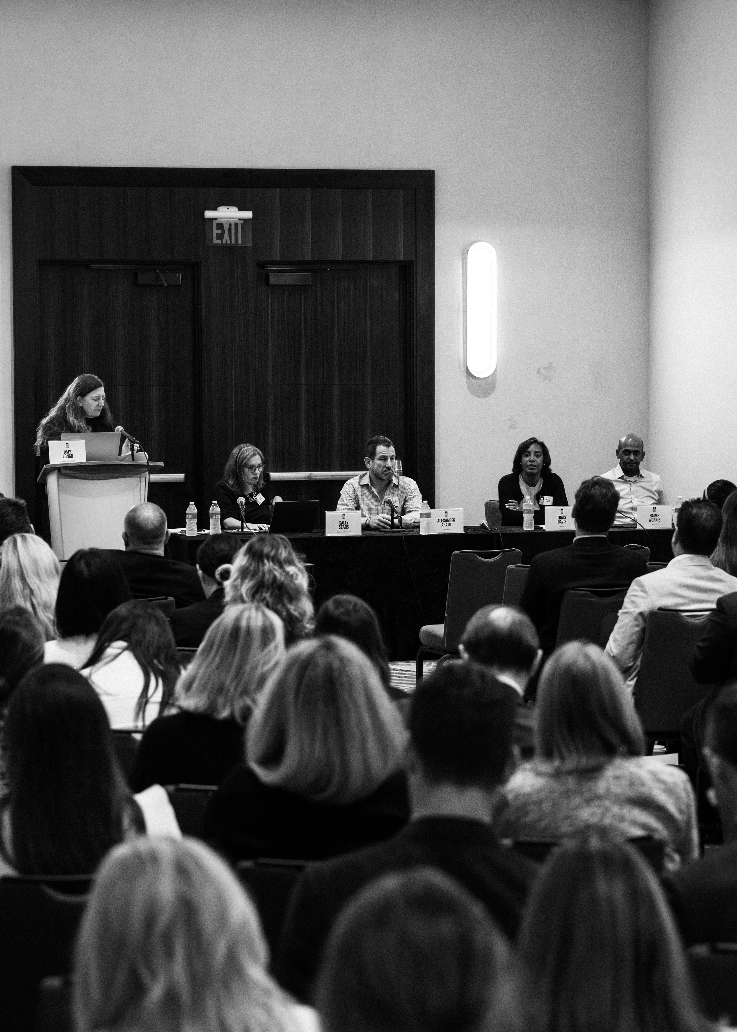 A panel of five people sitting at a long table with microphones and bottles of water, in front of an audience in a conference room. A woman is standing at a podium speaking.