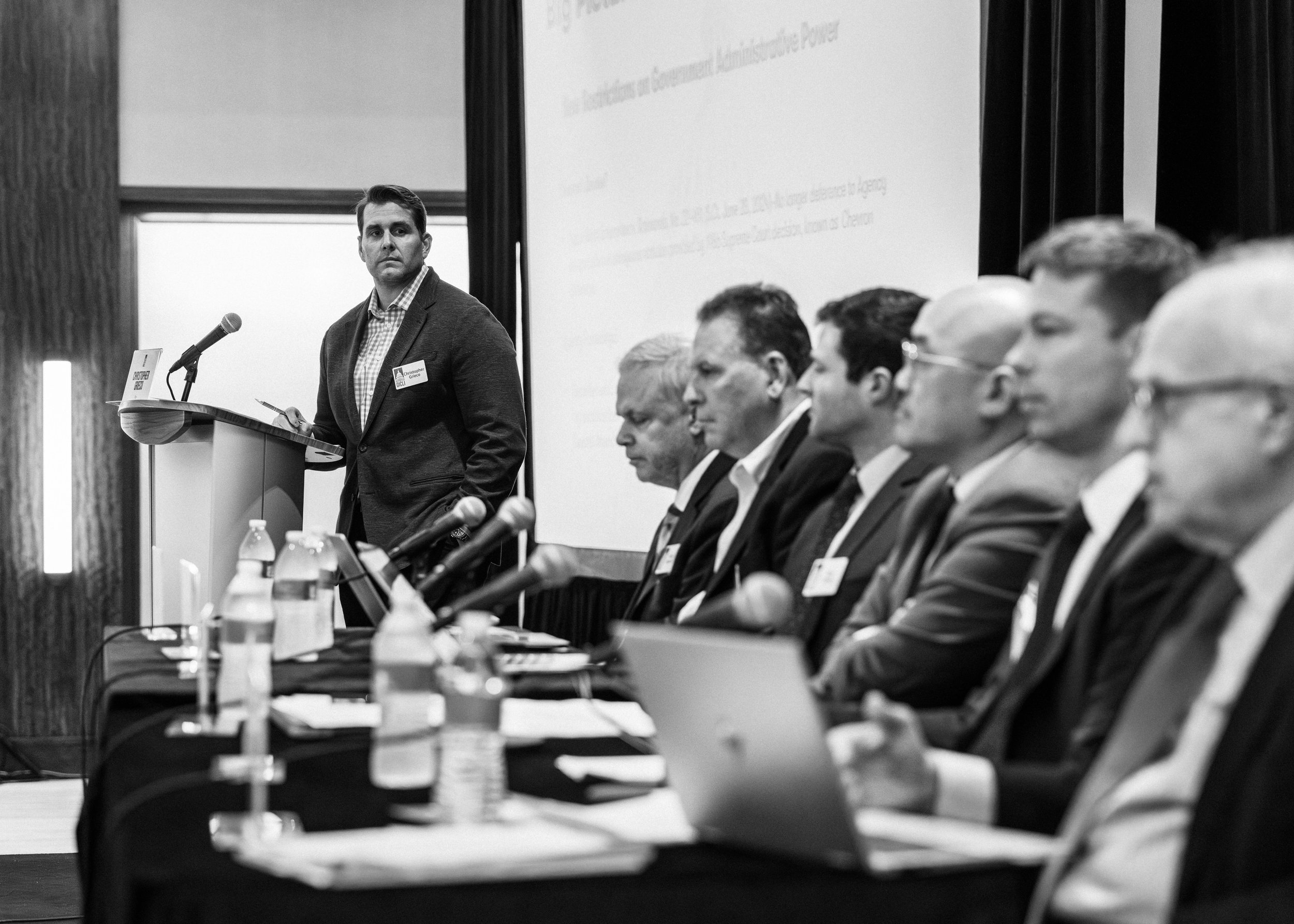 A black-and-white photo of a man standing at a podium with a microphone, looking towards a panel of seated men during a conference or meeting.