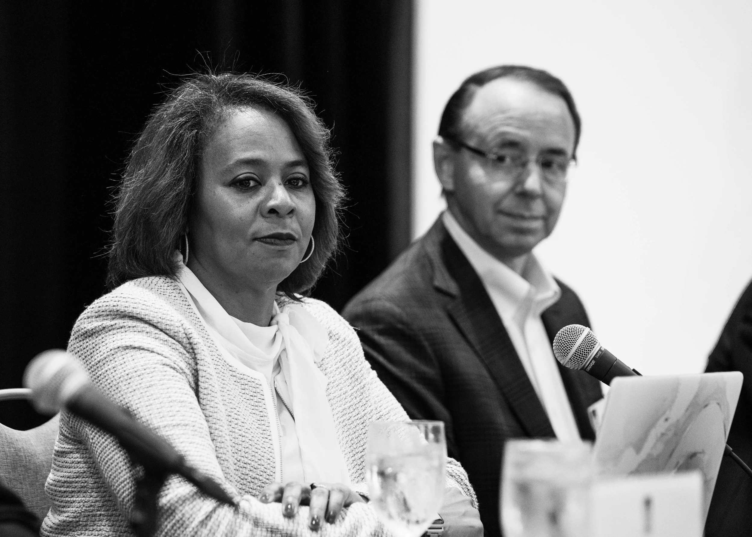 Black and white photo of a woman in a white blazer and a man in a suit sitting at a table during a panel discussion, with microphones and papers in front of them.