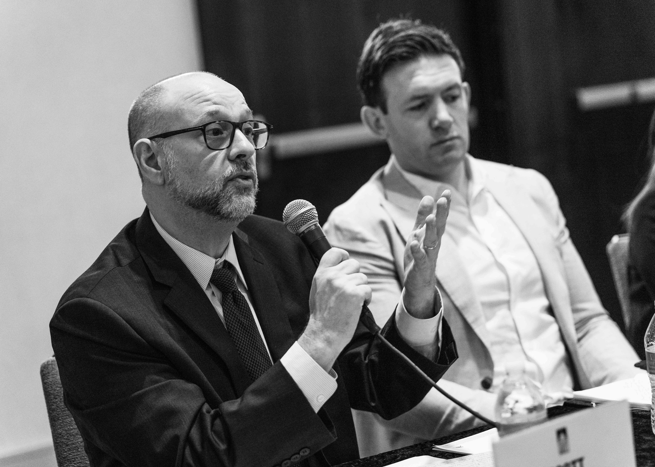 A man with glasses and a beard, dressed in a suit, speaking into a microphone during a panel discussion. Another man in a light suit jacket listens attentively.