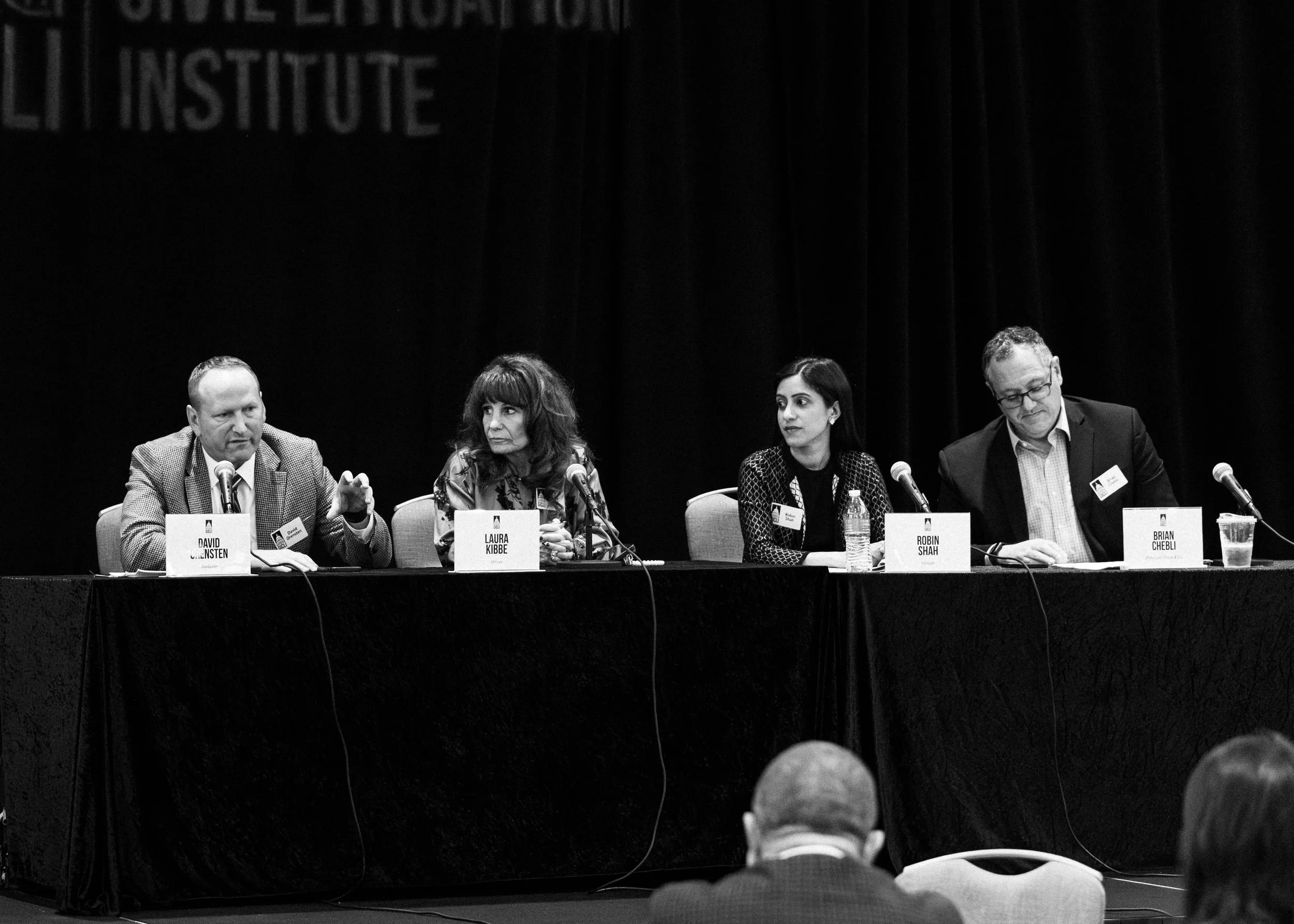 A panel of five people sitting at a long table at a conference, each with a nameplate and microphone. From left to right: David Klosensten, Laura Kibbe, Robin Shah, and Brian Chebli. The background is a dark curtain with a partially visible banner.