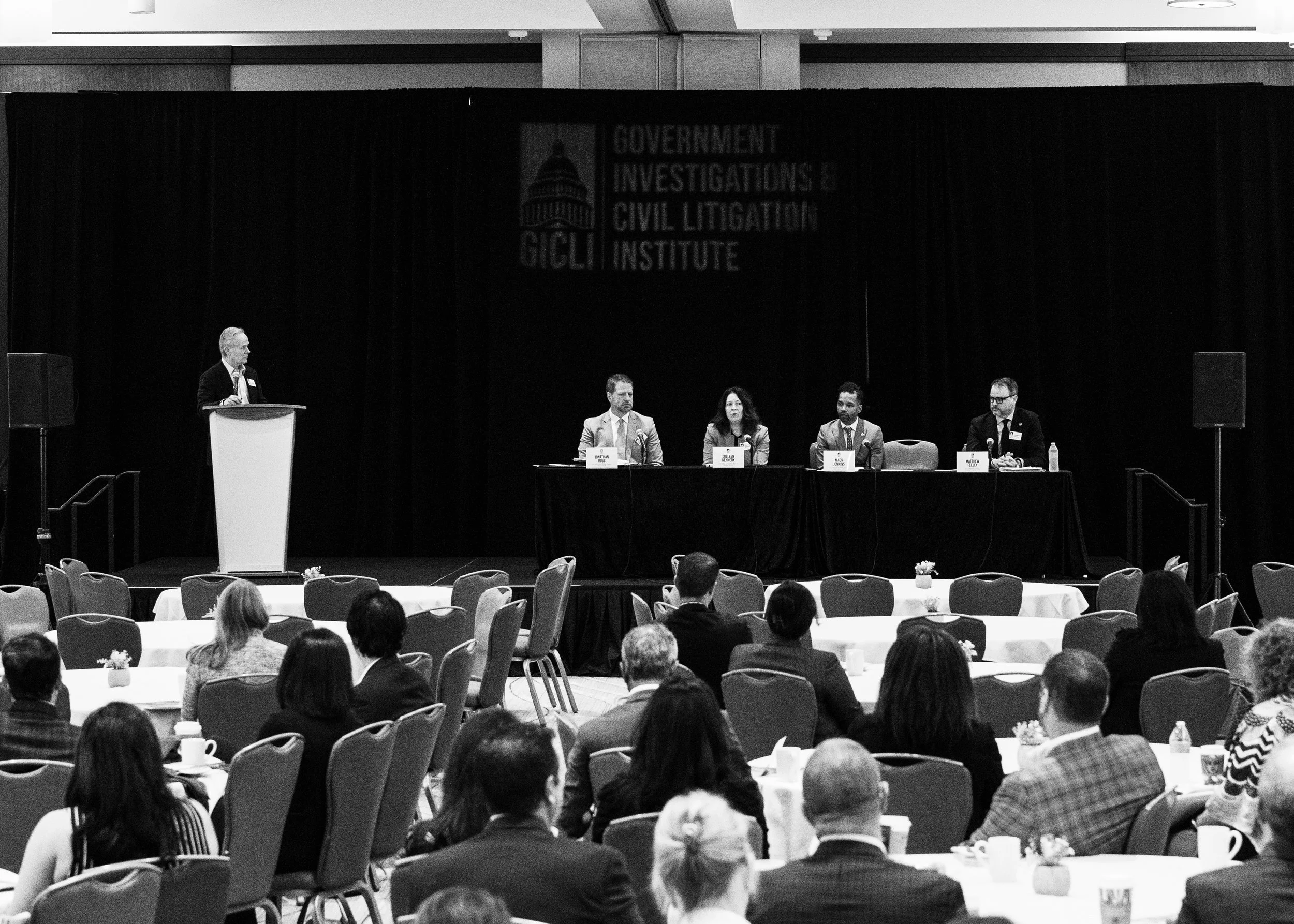 Panel discussion at a professional conference with audience seated at round tables, a speaker at a podium on stage, and a black curtain backdrop displaying the logo of the Government Investigations & Civil Litigation Institute.