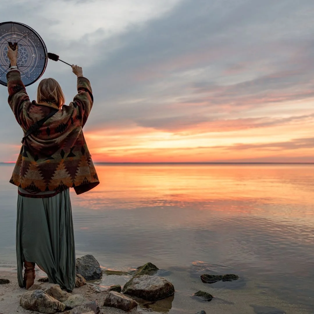 Person standing on rocky shore at sunset, holding a dreamcatcher with a butterfly inside, overlooking a calm body of water.