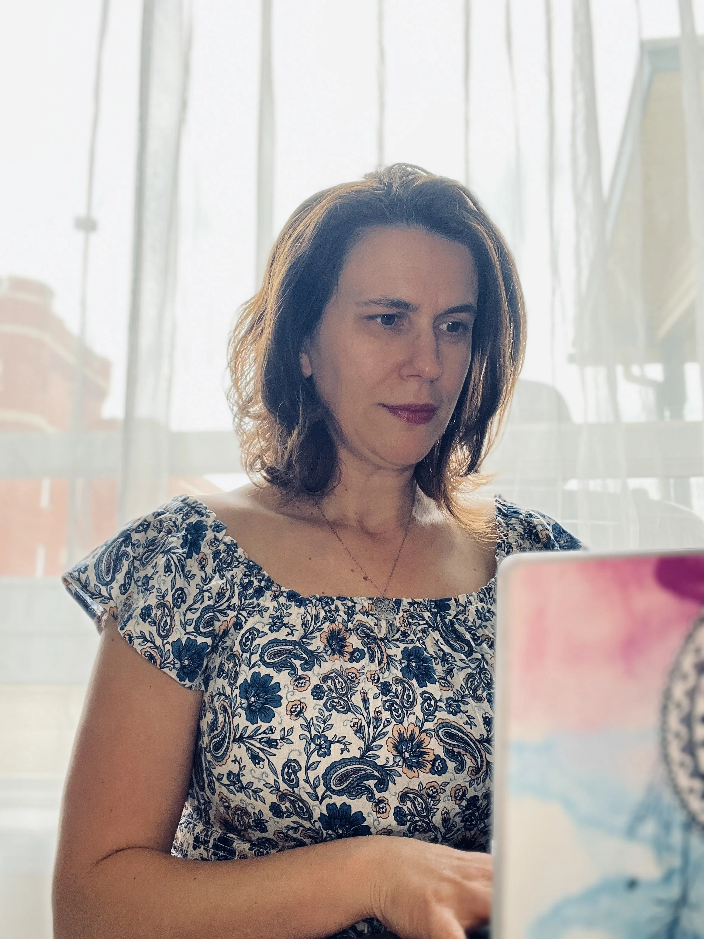 A woman with shoulder-length brown hair wearing a blue and white floral dress, holding a tri-coloured painting against a background of sheer curtains with sunlight streaming through.
