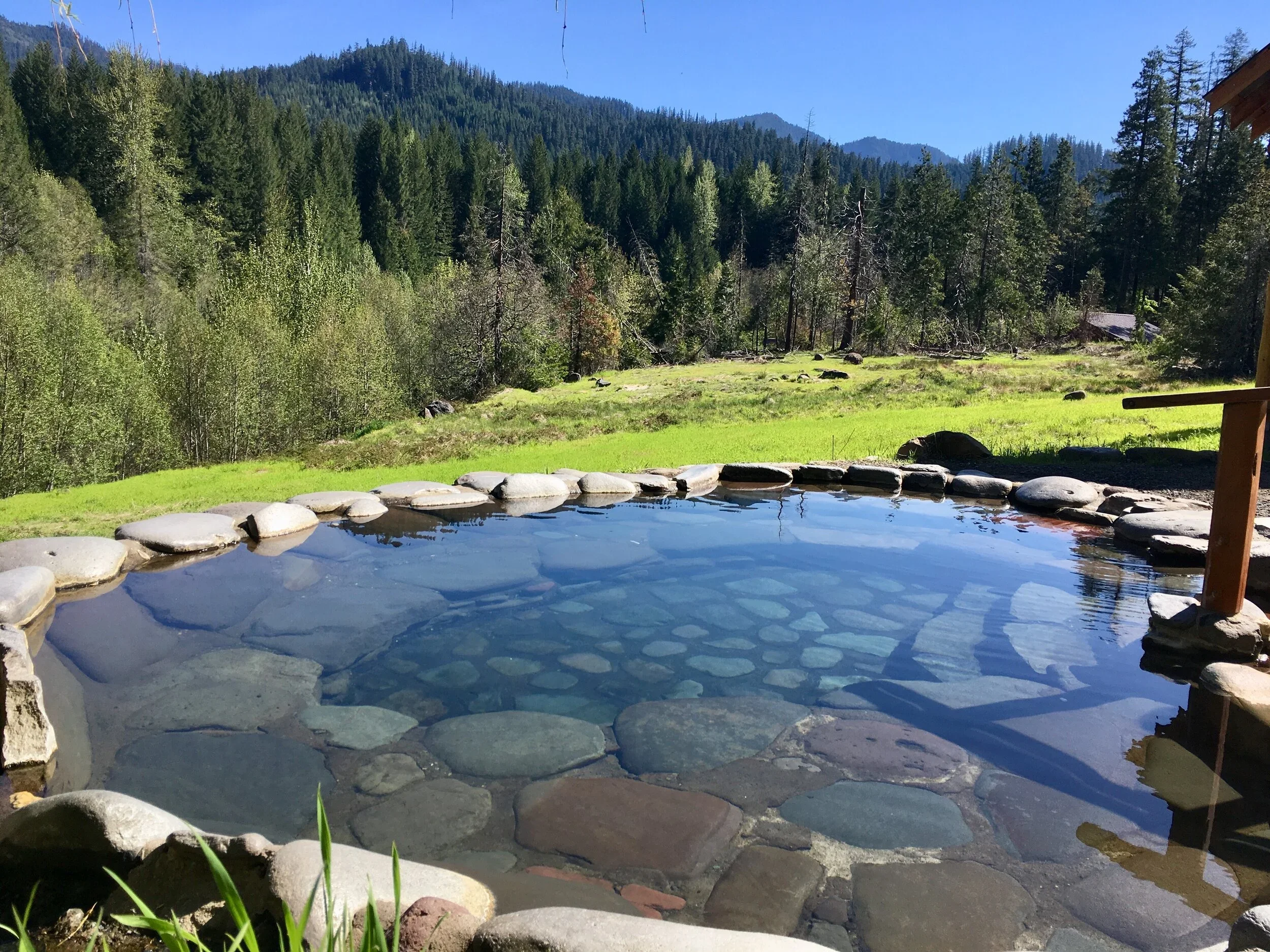 A natural hot spring pool with a stone border set in a lush green field, surrounded by forested mountains under a clear blue sky.