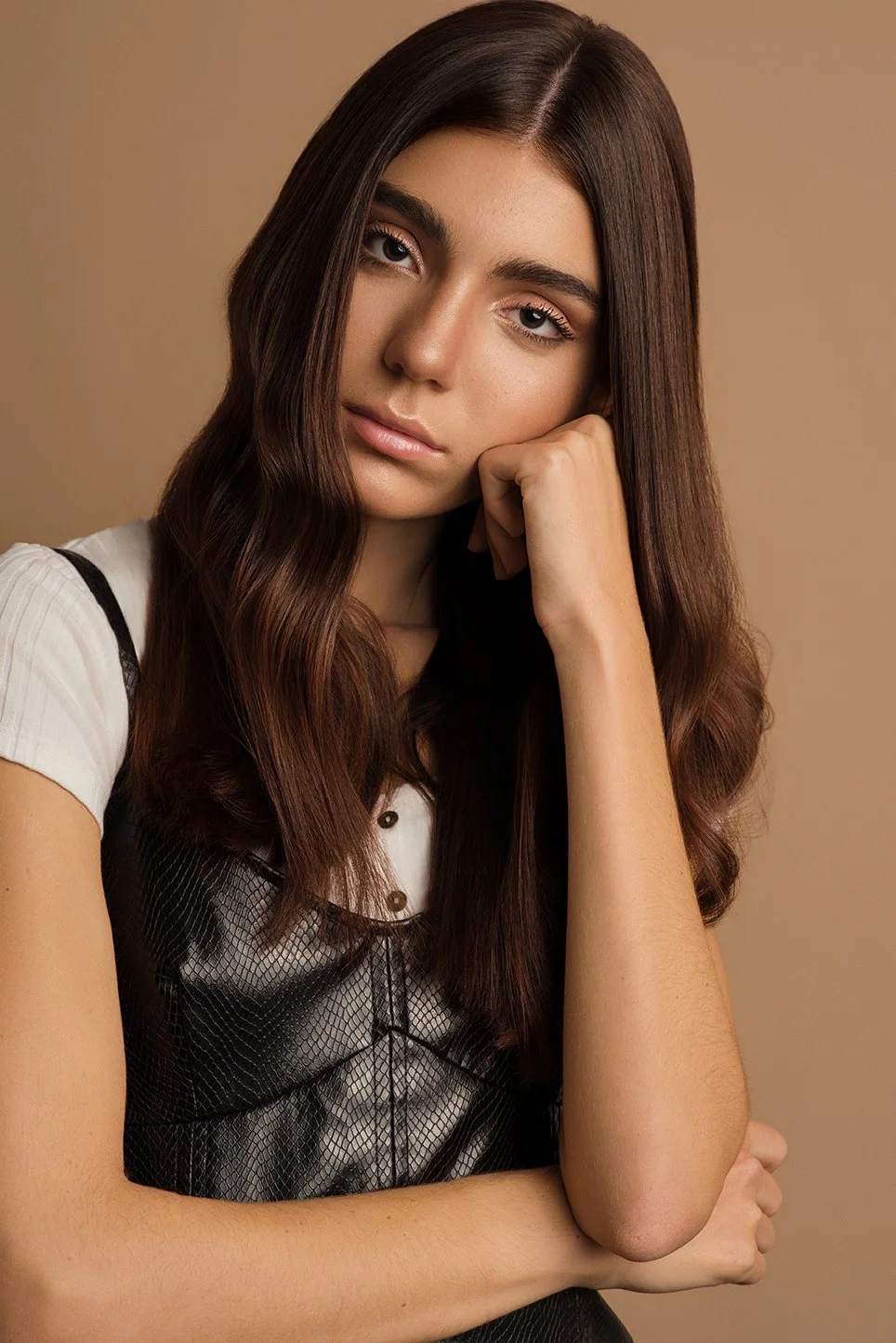 A young woman with long, wavy brown hair, resting her chin on her hand, wearing a white shirt and a black sleeveless top, posing against a beige background.