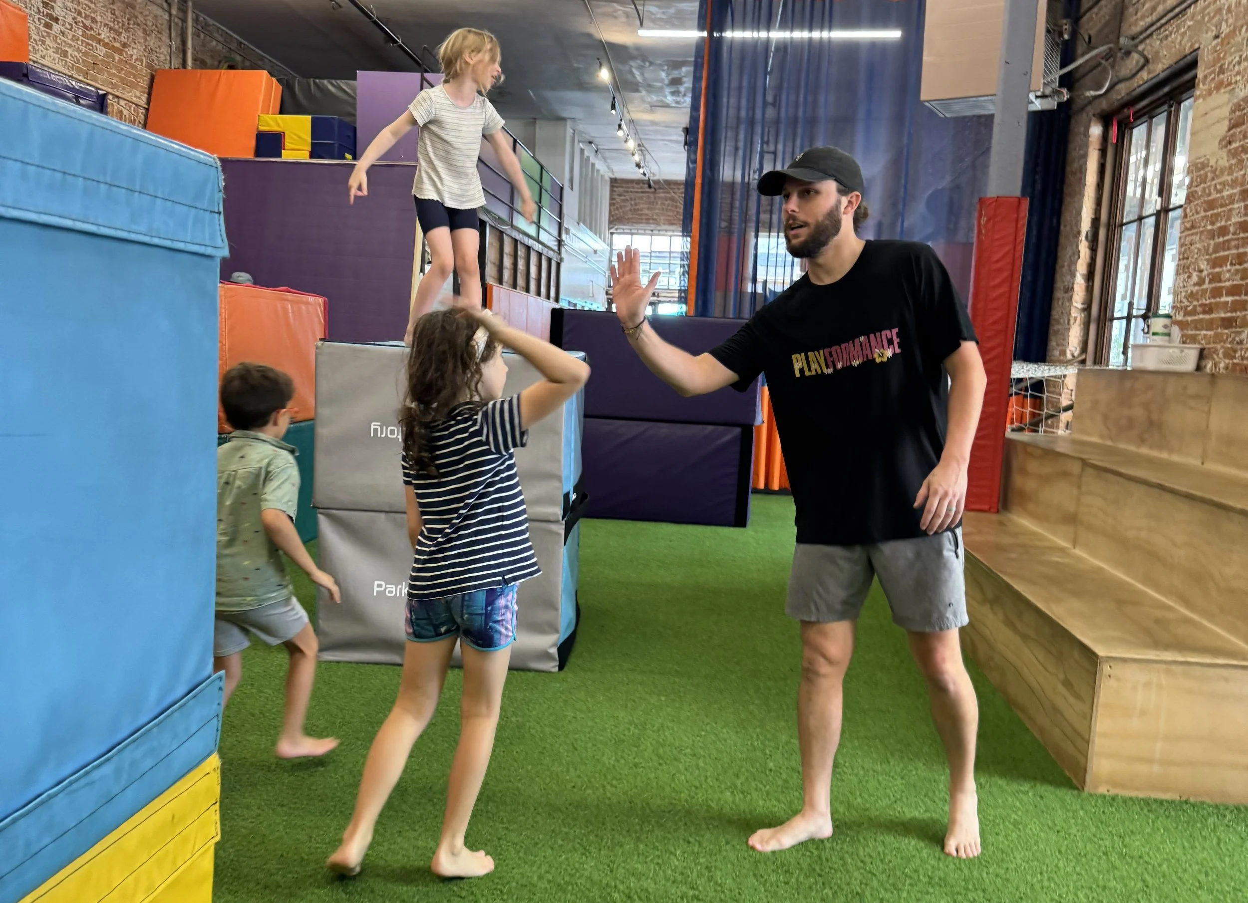 An adult man giving a high five to a young girl at an indoor trampoline park, with two younger children nearby, colorful padded walls and black netting in the background.