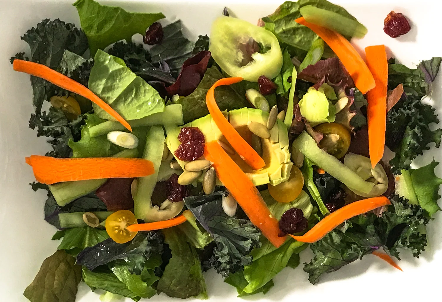 Composed salad of carrots, cucumbers, bell peppers and pepita seeds with tomatoes and cranberries on a bed of mixed greens with chopped kale