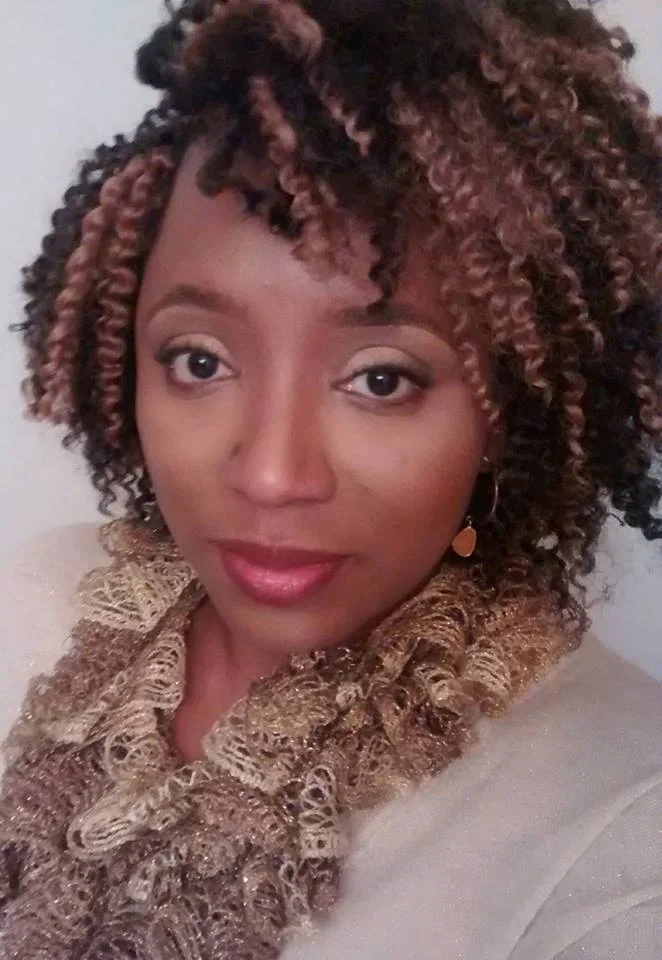 A woman with curly, two-toned hair smiling at the camera, wearing a gold lace scarf and gold earrings.