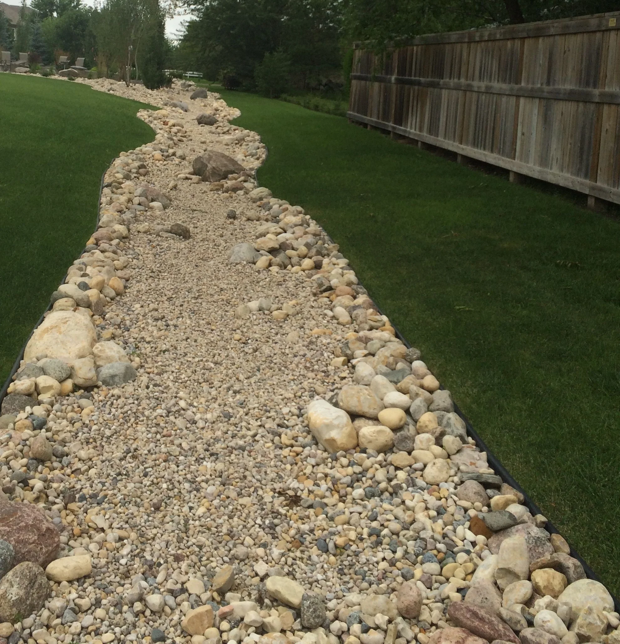 A landscaped backyard with a gravel pathway bordered by rocks, green grass, a wooden fence, and trees in the background.