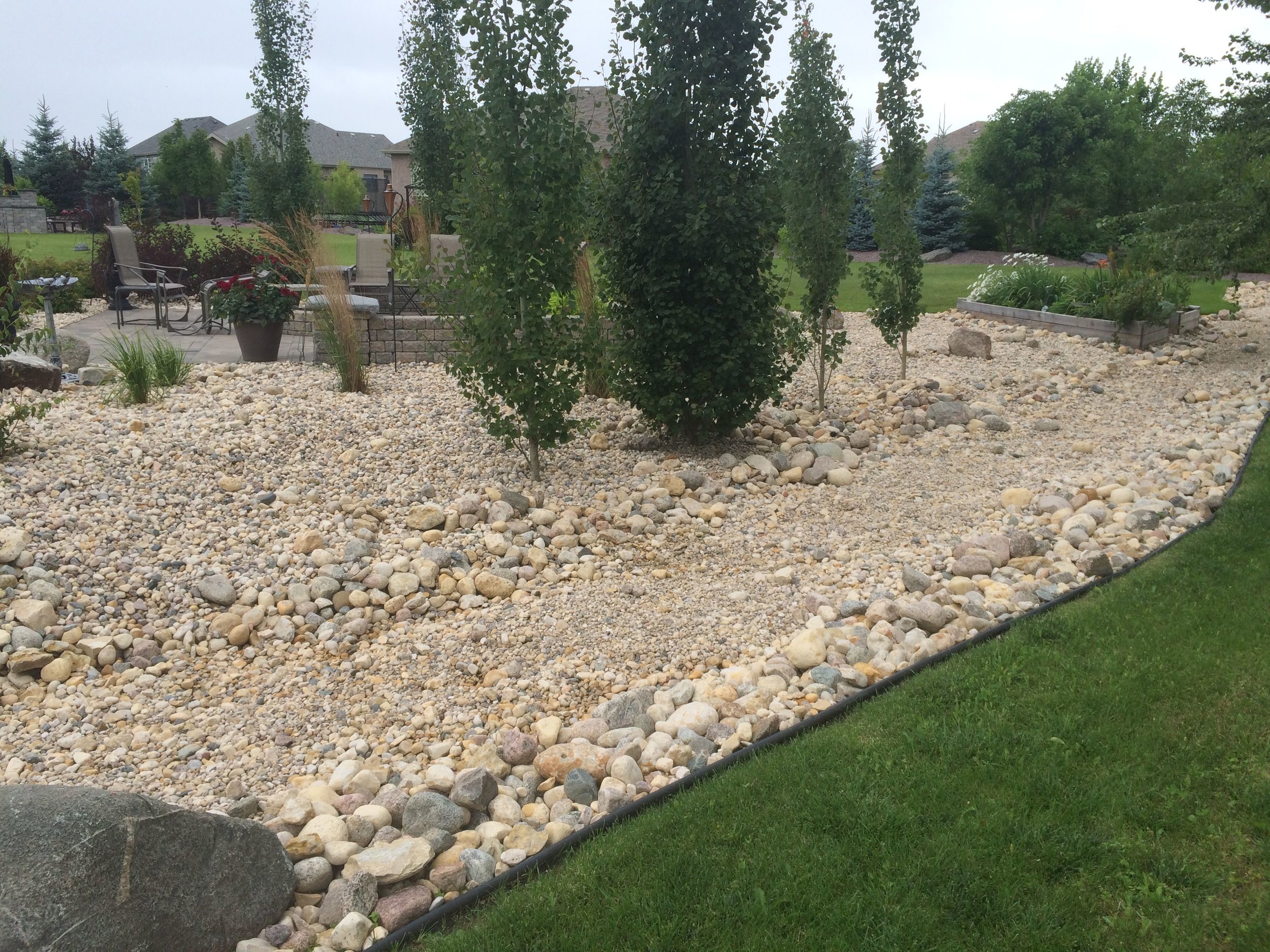 Residential backyard with rock landscaping, small trees, potted plants, outdoor seating, and a lawn area.