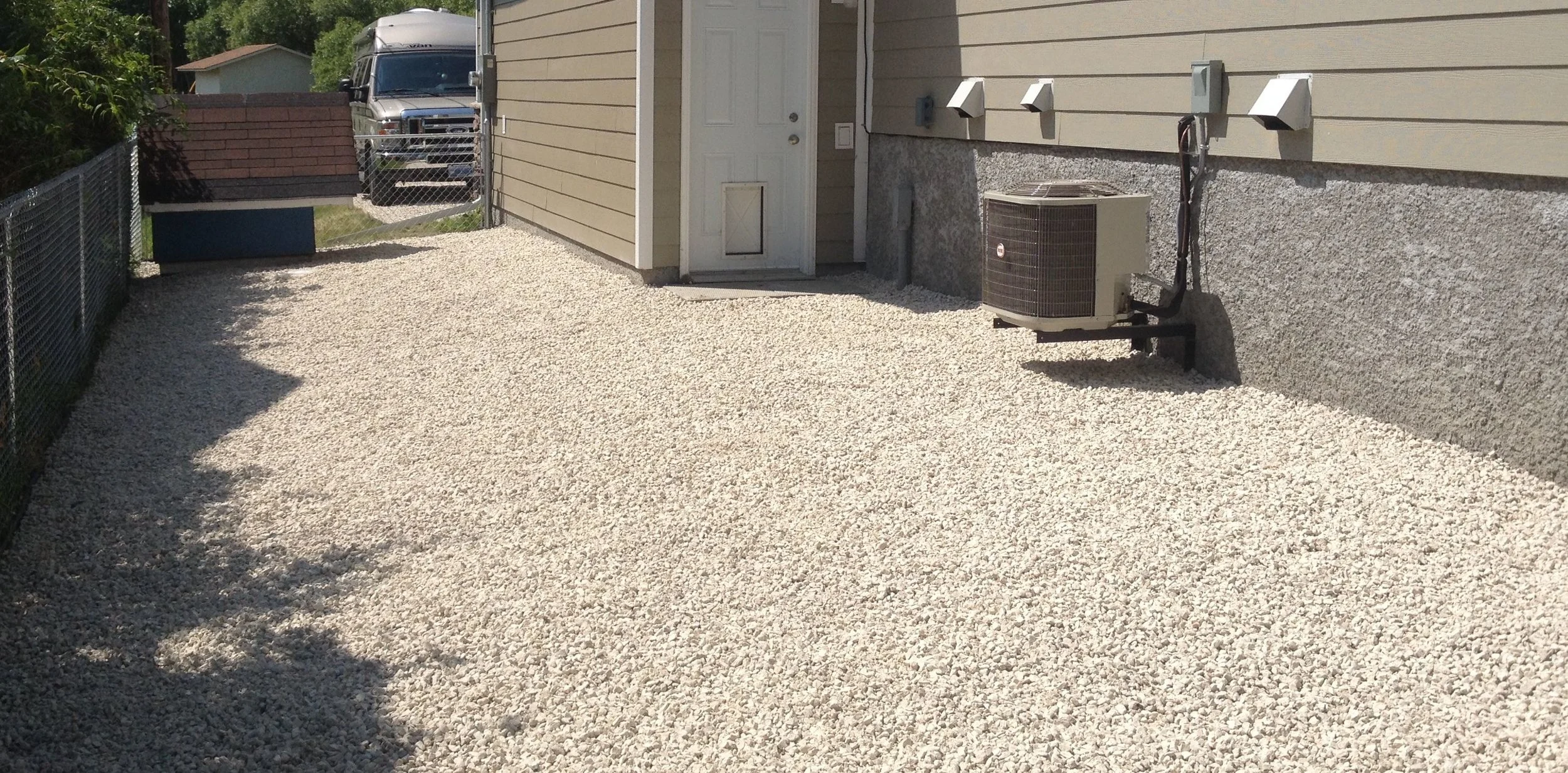 A backyard area with a gravel ground, enclosed by a wire fence on the left and the house wall on the right. The house has beige siding, a white door, and an air conditioning unit mounted on the wall. In the background, there are trees, a blue box, an
