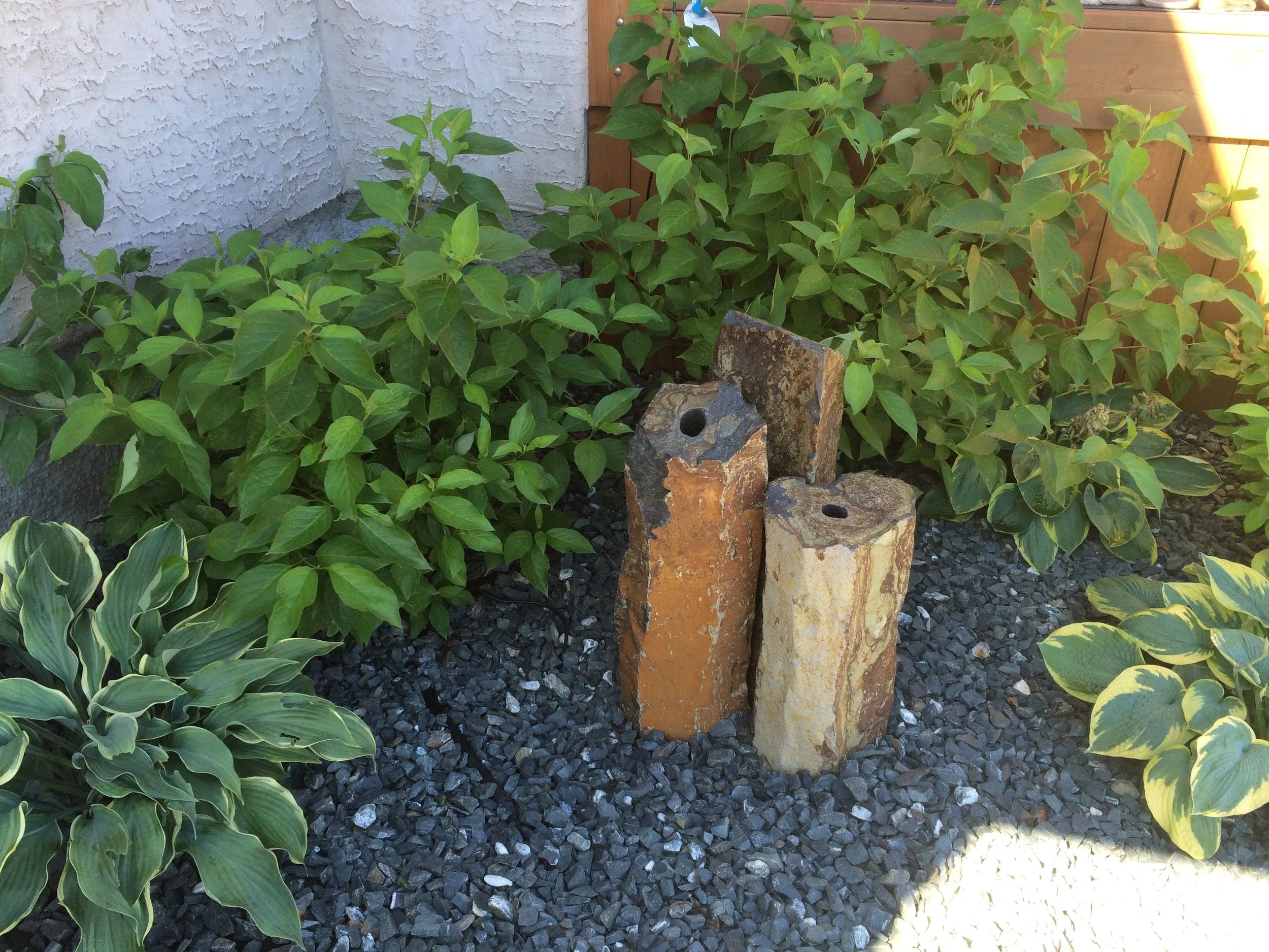 A garden corner with green leafy plants, black gravel ground cover, and three irregular-shaped stone sculptures with holes in the center, set against a wooden fence and a white textured wall.
