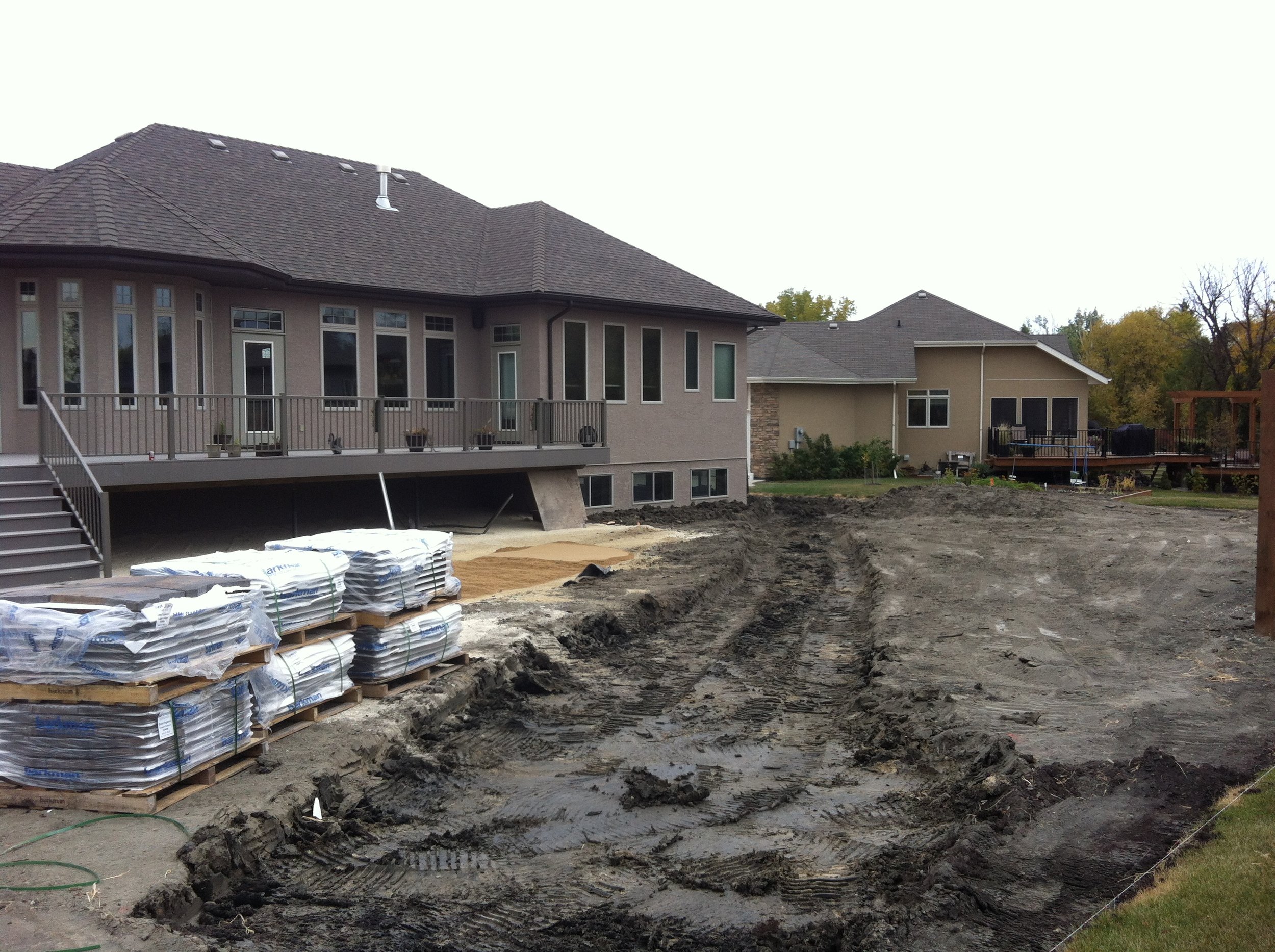 Backyard under construction with dirt and tire tracks, stacked bags of construction material in the foreground, and houses with decks in the background.