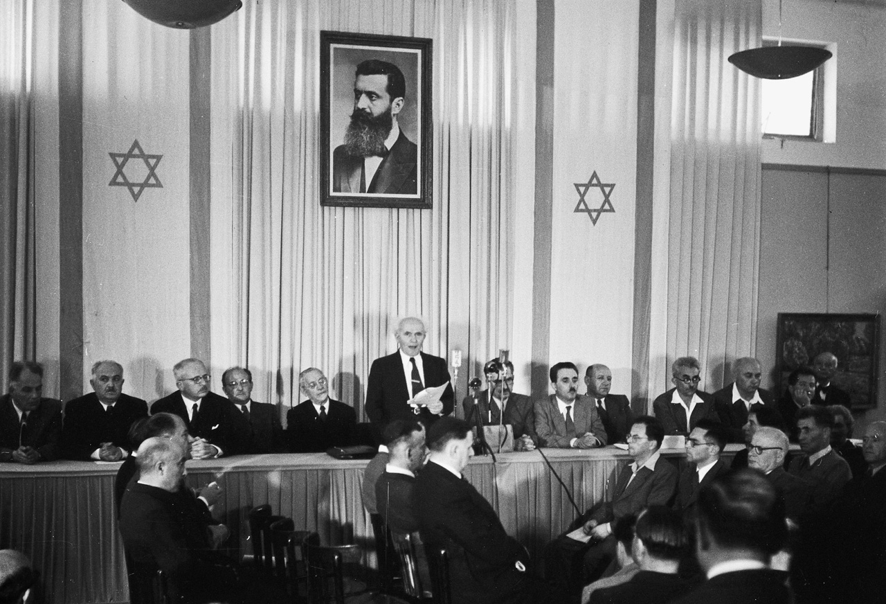 David Ben-Gurion, who engineered the founding of Israel and was its first Prime Minister, declaring independence beneath a portrait of Theodor Hertzl, the father of Zionism.